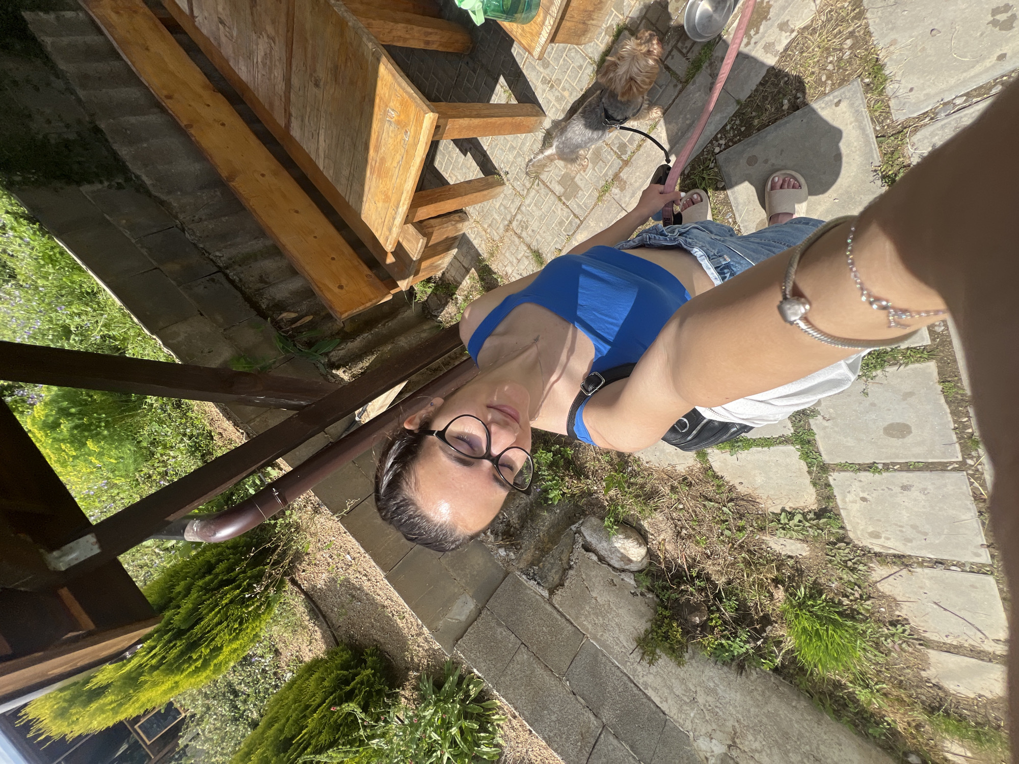 The image captures a selfie-style scene featuring a young woman and a small dog in an outdoor environment in Alino, Bulgaria. The woman, appearing to be in her 20s to early 30s, has dark hair pulled back and wears glasses, a blue crop top, denim shorts, and light-colored sandals. She is holding the camera high, with her right arm and a silver bracelet visible in the immediate foreground, obscuring part of her lower body. She smiles with her eyes partially closed. A black bag strap is visible over her left shoulder.
To her right, a small dog, consistent with a Yorkshire Terrier with brown and grey fur, stands on a paved surface. The dog is on a pink leash, which appears to be held by the woman. Near the dog, a silver pet bowl rests on the ground. The ground consists of grey square concrete paving slabs, with grass and weeds growing between the gaps.
The background features a wooden outdoor dining set, comprising a table and benches, made of light-toned wood. Above and to the left, part of a dark brown wooden structure, likely a pergola or shelter, is visible with a brown drainage pipe attached. A concrete block retaining wall separates the paved area from an elevated, green landscaped section. This section contains several tall, conical evergreen shrubs and various green plants, some with small purple flowers. Strong sunlight illuminates the scene, casting distinct shadows. A green plastic bottle is visible on the wooden table.