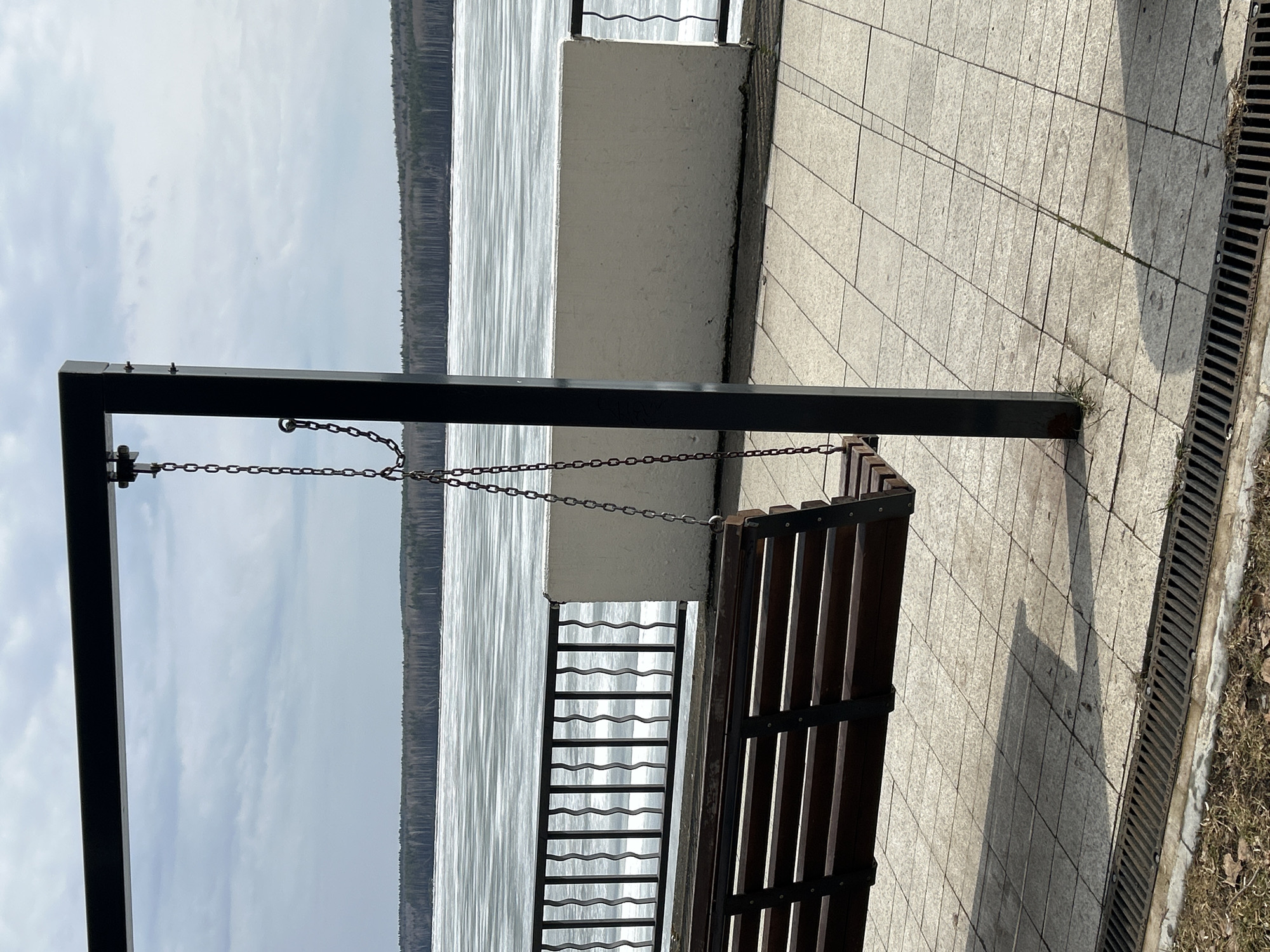 A swing bench overlooks the Kama River in Chaikovsky, Russia. The bench, suspended by chains from a black metal frame, sits on a paved walkway near a drainage grate. Beyond a short, white wall and metal railing, the river stretches towards the horizon, with forested banks visible in the distance. The sky is overcast.