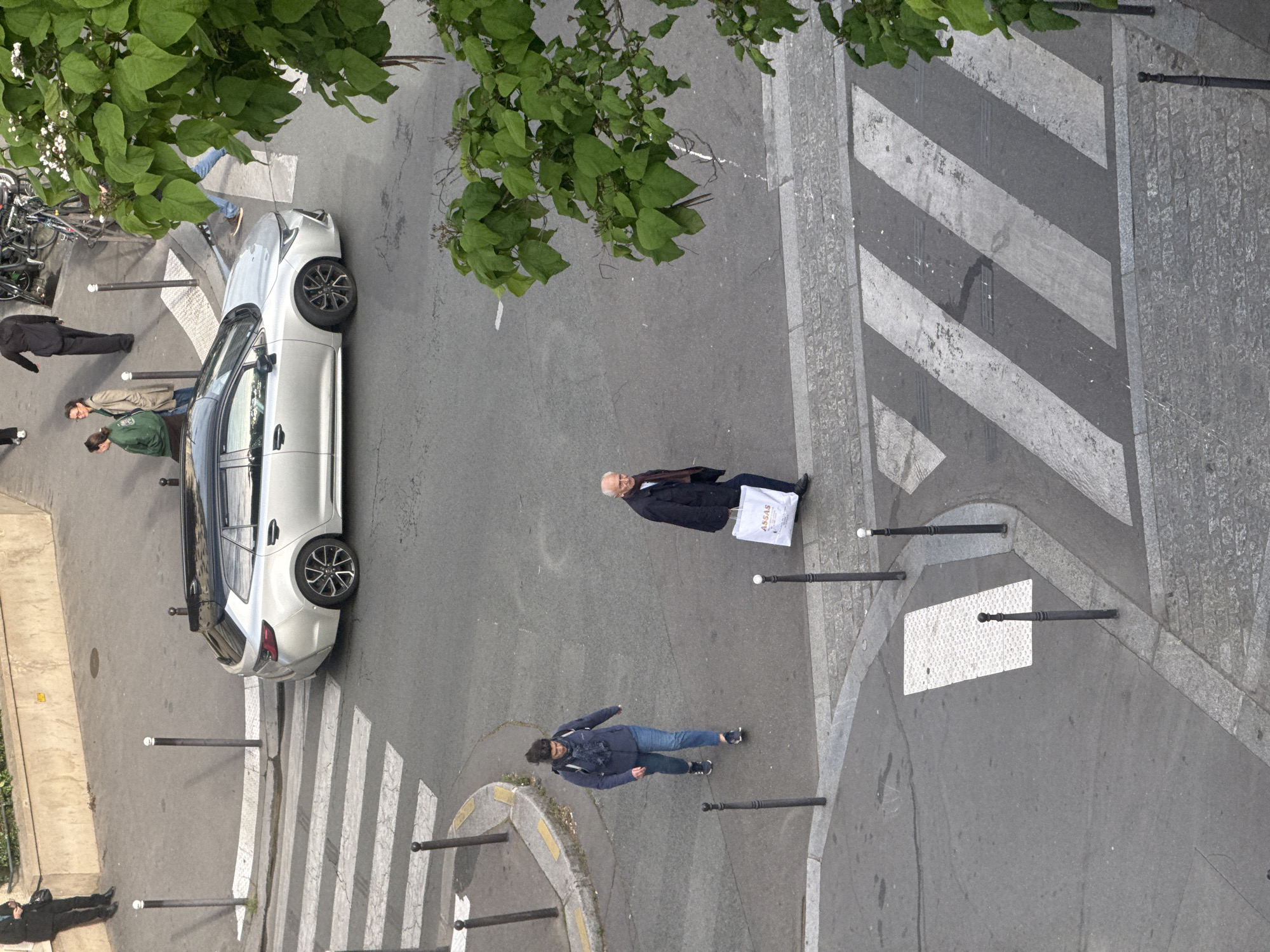 An aerial view in Paris, France, showcases a street scene featuring several people, a parked car, and street furniture. A white car is parked alongside a crosswalk. Two individuals are seen walking on the sidewalk, one in a suit carrying a shopping bag. A woman is walking on the street edge. Other pedestrians are visible along the sidewalk. The scene is partially shaded by trees. Metal bollards line the sidewalk. A bicycle rack with parked bikes is also visible. Crosswalks, street corners, and a cobblestone road contribute to the Parisian setting.