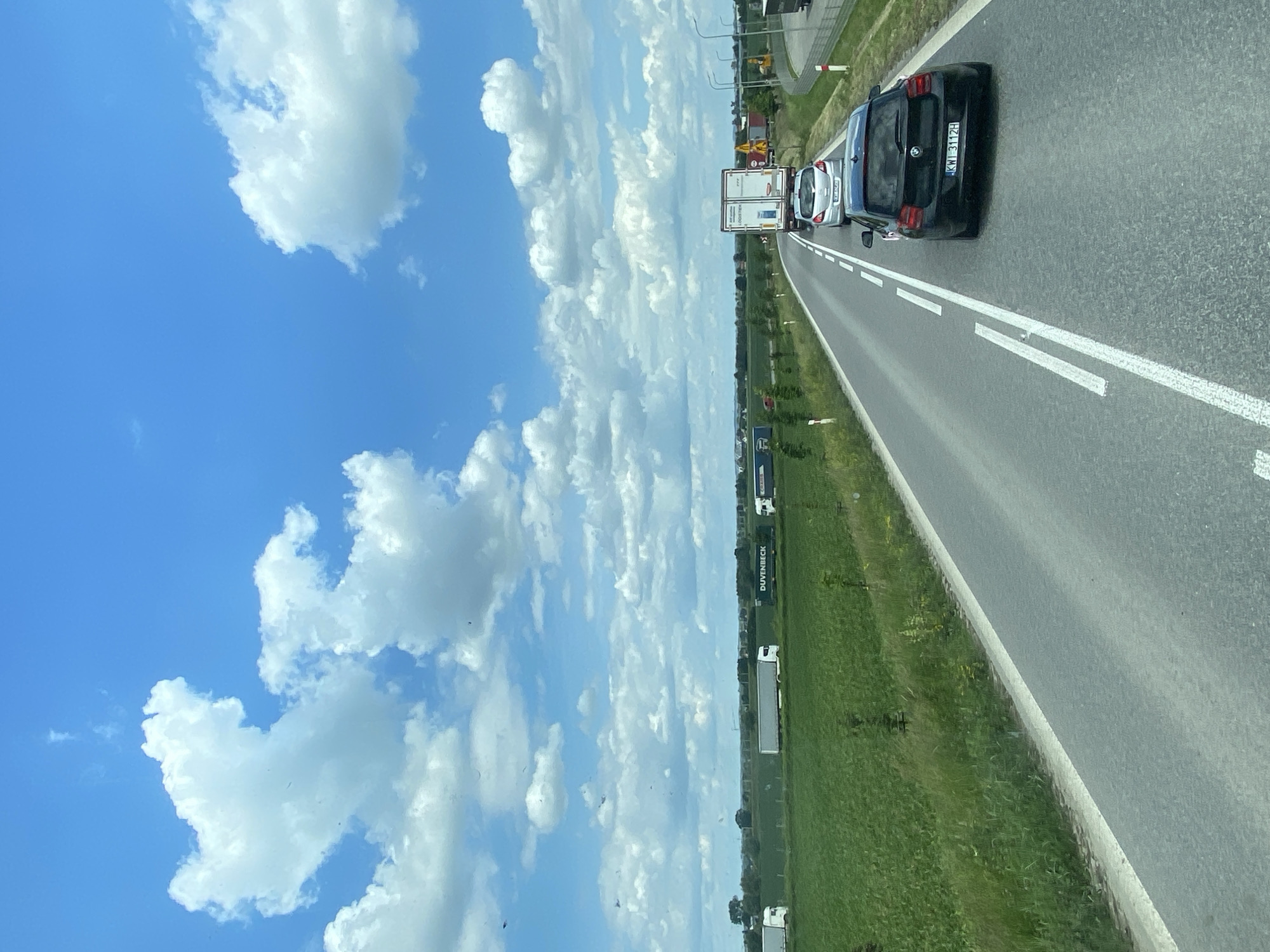 In Podłęże, Poland, on a sunny day with a bright blue sky dotted with fluffy white clouds, a traffic jam is visible on a highway. The road features white lane markings. In the foreground, a black BMW hatchback with license plate "KWI 3112H" is stationary, followed by a silver hatchback and a white truck. Beyond the road, a vast green field stretches towards the horizon, with several white and green trucks and trailers parked along the roadside in the distance. Trees are scattered throughout the landscape, and the overall scene suggests a rural or semi-rural setting.