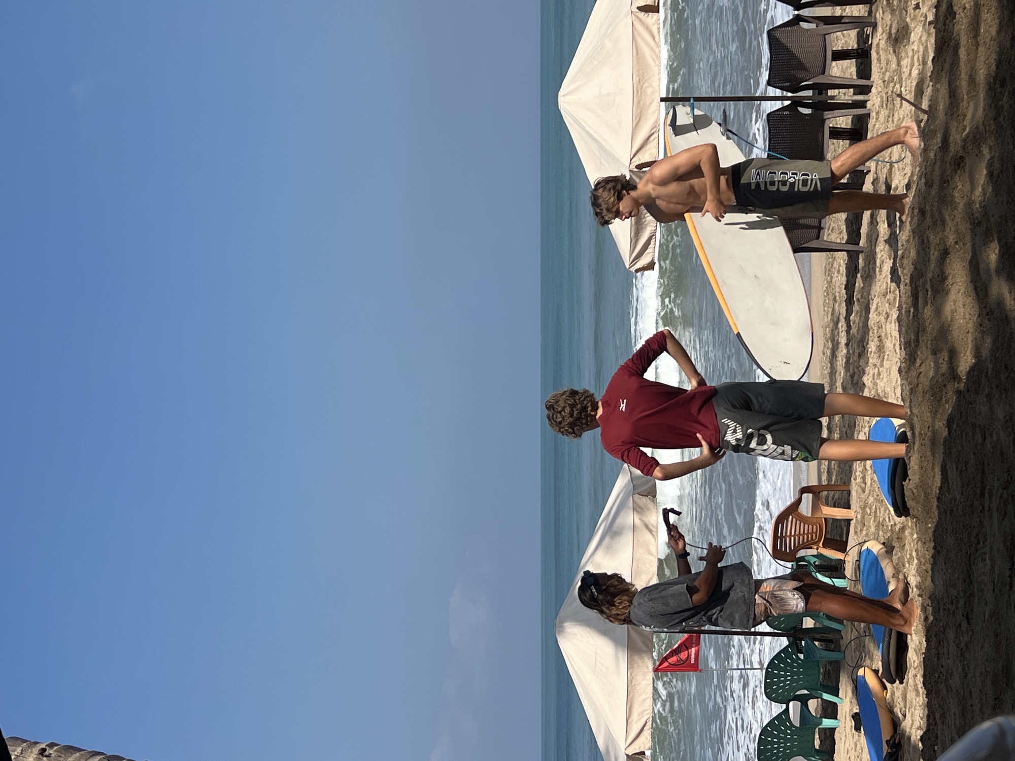Three individuals are on a sandy beach in Kuta, Indonesia. Two young men, one shirtless and wearing dark boardshorts with white text, and another wearing a dark red long-sleeved shirt and grey shorts, stand facing the ocean. The shirtless young man holds a white surfboard with an orange stripe. The other young man has his hands on his hips. A third individual, with long brown hair and wearing a grey t-shirt and patterned shorts, stands between them and slightly to the left. This person appears to be holding a black object with a cord attached, possibly a camera or a piece of surfing equipment.
Behind the people, there are two large, cream-colored beach umbrellas, one slightly angled towards the ocean. Several plastic chairs, some green and some brown, are visible in front of the umbrellas. A red flag with white markings is planted in the sand near the left umbrella. In the foreground, several blue and yellow surfboards are lying flat on the sand. The ocean is visible in the background, with waves breaking on the shore. The sky is clear and blue, indicating a sunny day. The sand is light brown, and shadows are cast on the ground, suggesting the sun is high.