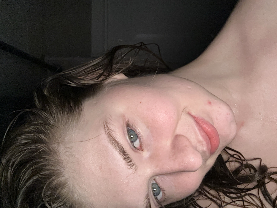 A close-up of a person's face and upper chest, rotated sideways. The person has wet, dark hair and light skin. Their eyes are blue. There's a darkened background visible above and to the side of their head, and a small birthmark is visible on the nose. Water droplets glisten on the skin, and the expression is a slight smile.