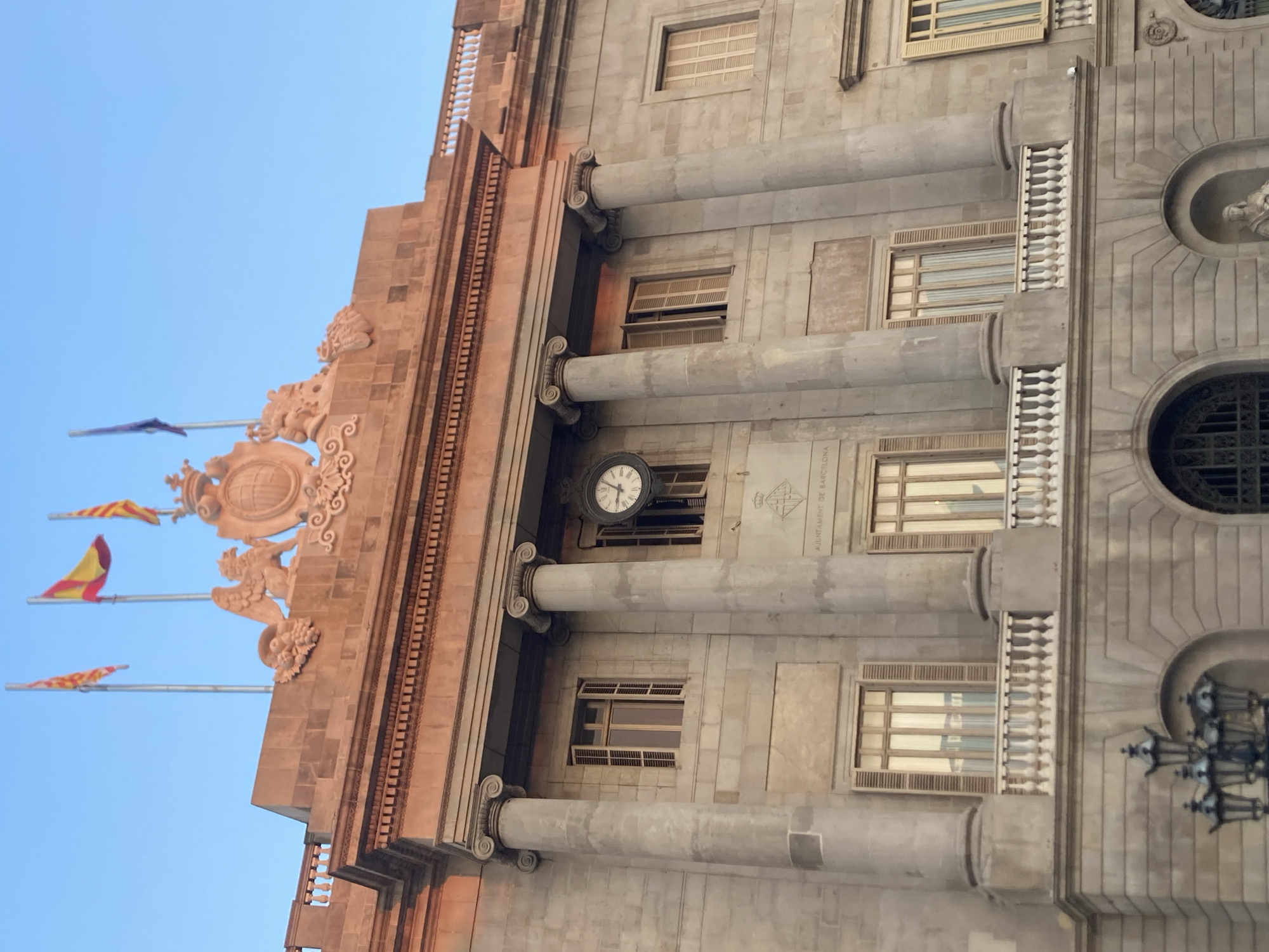 The image shows the Ajuntament de Barcelona, or City Hall, located in Barcelona, Spain. The facade of the building is made of stone and features classical architectural elements such as columns, arches, and balconies. There are several windows on the building, some with wooden shutters. A prominent clock is mounted on the facade. Above the building, three flags are visible against a clear blue sky. The building is illuminated by warm, reddish light, suggesting it is either sunrise or sunset. The overall impression is one of grandeur and historical significance.