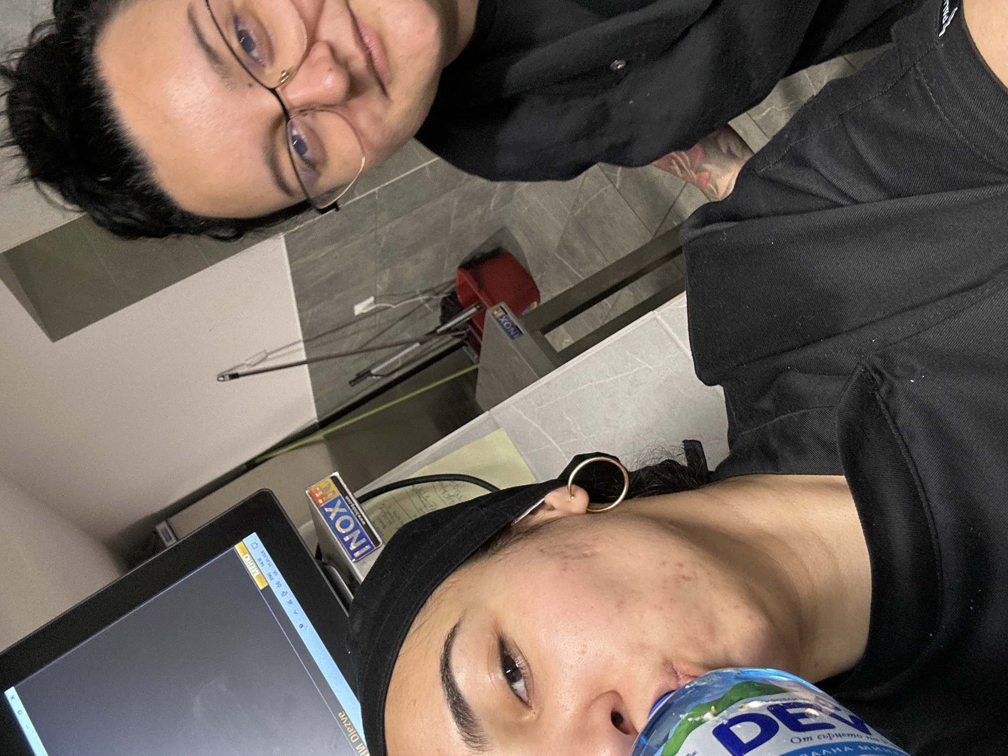 The image depicts two individuals in an indoor setting. One person is seen wearing glasses and a black shirt. A visible tattoo is on their arm. The second person is wearing a black cap and a black shirt, and a gold hoop earring is visible. They are drinking from a bottle of "Devin" water. A computer monitor is present on a table. A red bucket labelled "INOX" is also visible in the background. The inscription on the water bottle is in Bulgarian: "От сърцето на Родопите" (“From the heart of the Rhodopes”). Burgas is not specifically apparent, though the water bottle's branding could suggest a Bulgarian location.