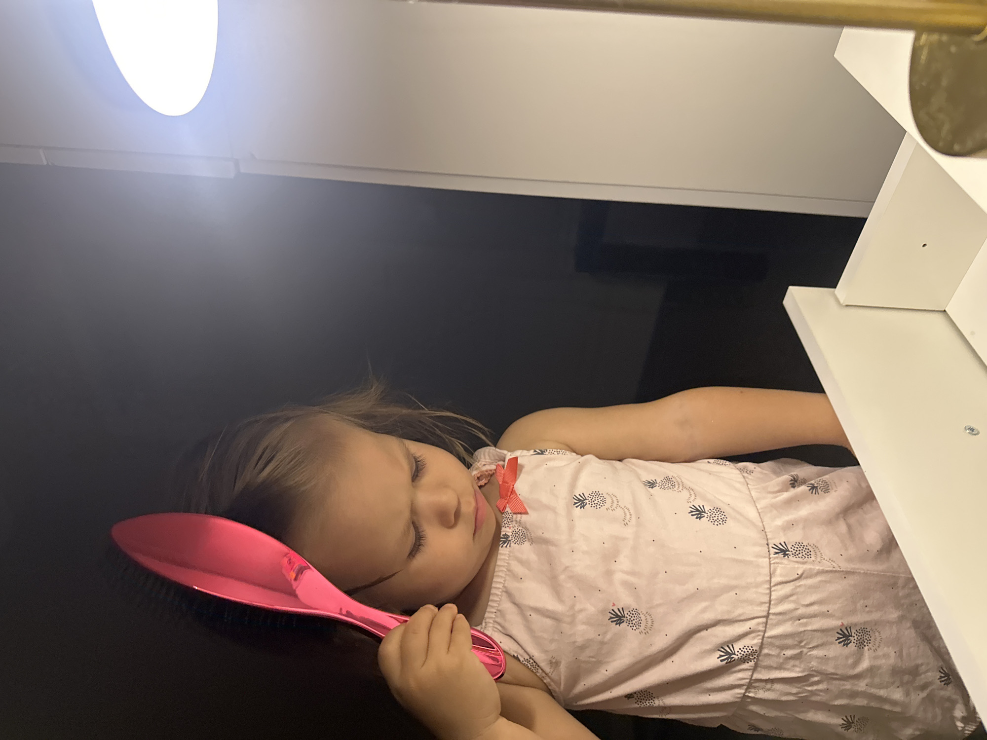 A young child with blonde hair, wearing a light pink romper with a pineapple pattern and an orange bow, is holding a bright pink hairbrush over their head. The child is looking down with a pensive expression. The scene appears to be indoors, with a bright light source to the right, casting shadows and illuminating part of a white surface in the foreground, possibly furniture. The background is dark. The specific location within Kharnavy, Russia, is not determinable from the image.