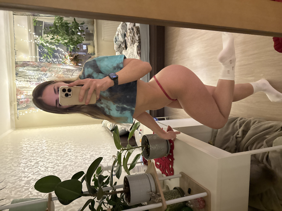 A woman takes a mirror selfie. She is wearing a tie-dye crop top, a red thong, and white socks. She's holding her phone in her left hand, showing off her watch. Her brown hair hangs loosely. In the background, there is a shelf with plants, a window with fairy lights, and a bed.