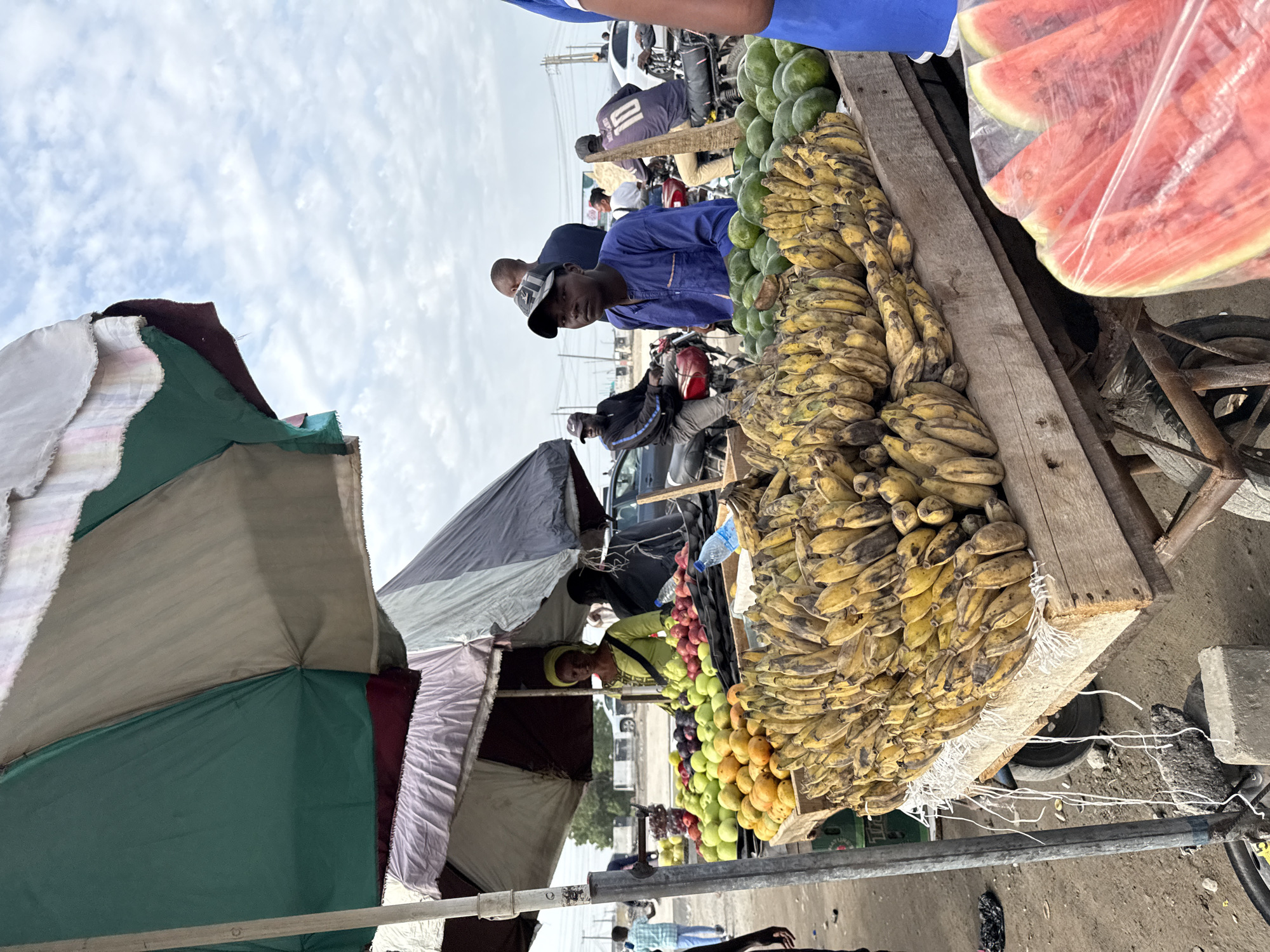 The image captures a vibrant street market scene in Lekki, Nigeria. A vendor in a blue shirt and cap stands behind a wooden cart piled high with ripe bananas. Next to the bananas are piles of green mangos. Above the bananas and mangoes is a stack of plastic wrapped watermelon slices. The cart's wheels are visible, suggesting it's mobile. Other vendors and customers are seen in the background, some on motorcycles. Overlapping umbrellas of various colors provide shade to the market stalls. The sky overhead is overcast.