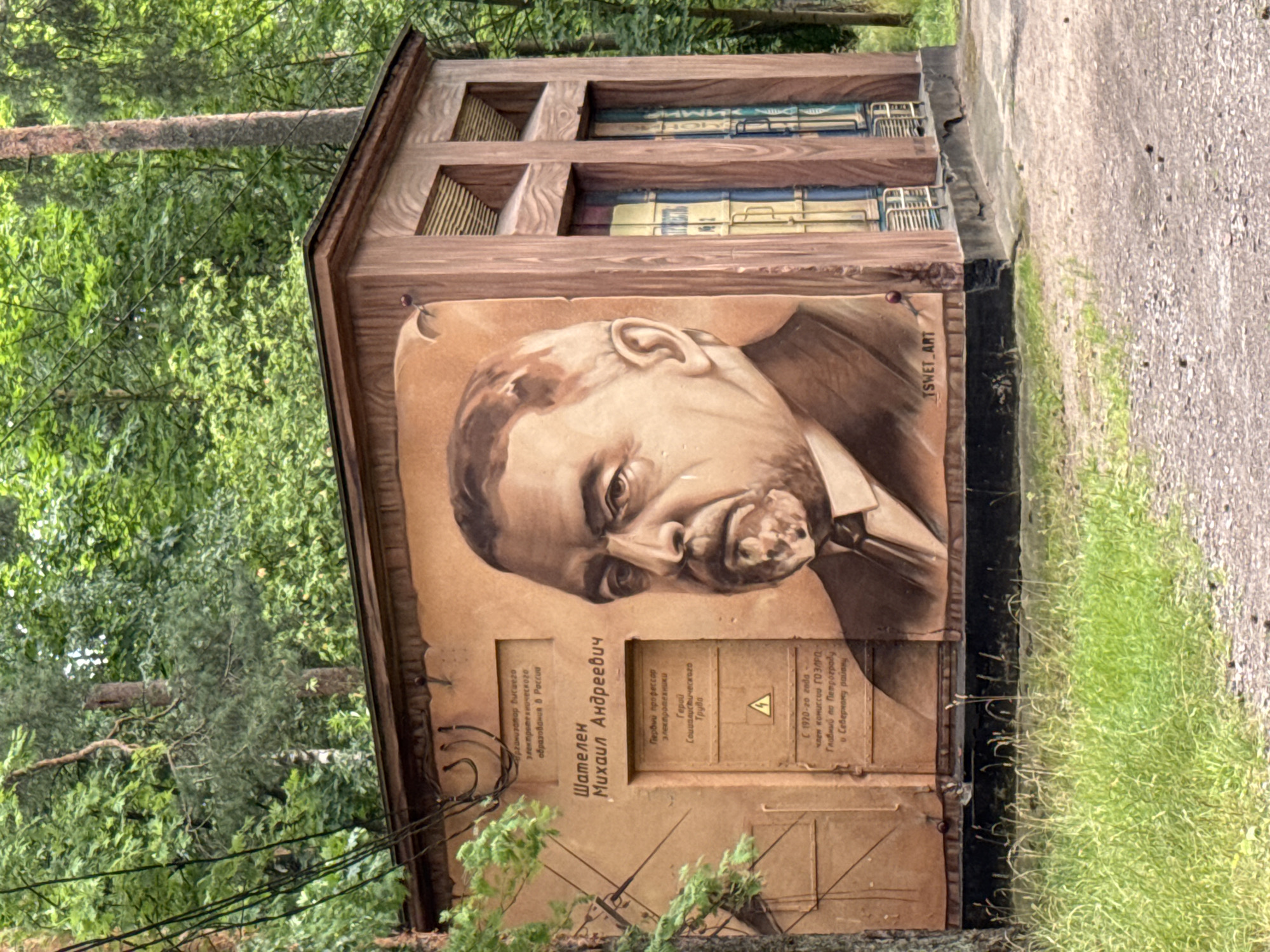 The image depicts a mural of a man's portrait on the side of a wooden utility building located in Ladozhskoe Ozero, Russia. The mural features a large, realistic painting of a man with a stern expression, likely a historical or important figure. To the left of the portrait, there is text in Cyrillic, and below it, what appears to be a door or panel with more Cyrillic text and a caution symbol. The building itself is made of wood and has a sloped roof. It is situated in an outdoor environment with lush green trees and foliage surrounding it, suggesting it's in a park or wooded area. The ground in front of the building is covered with gravel and grass. The overall scene is static, with no visible human activity other than the presence of the mural as a form of art installation.