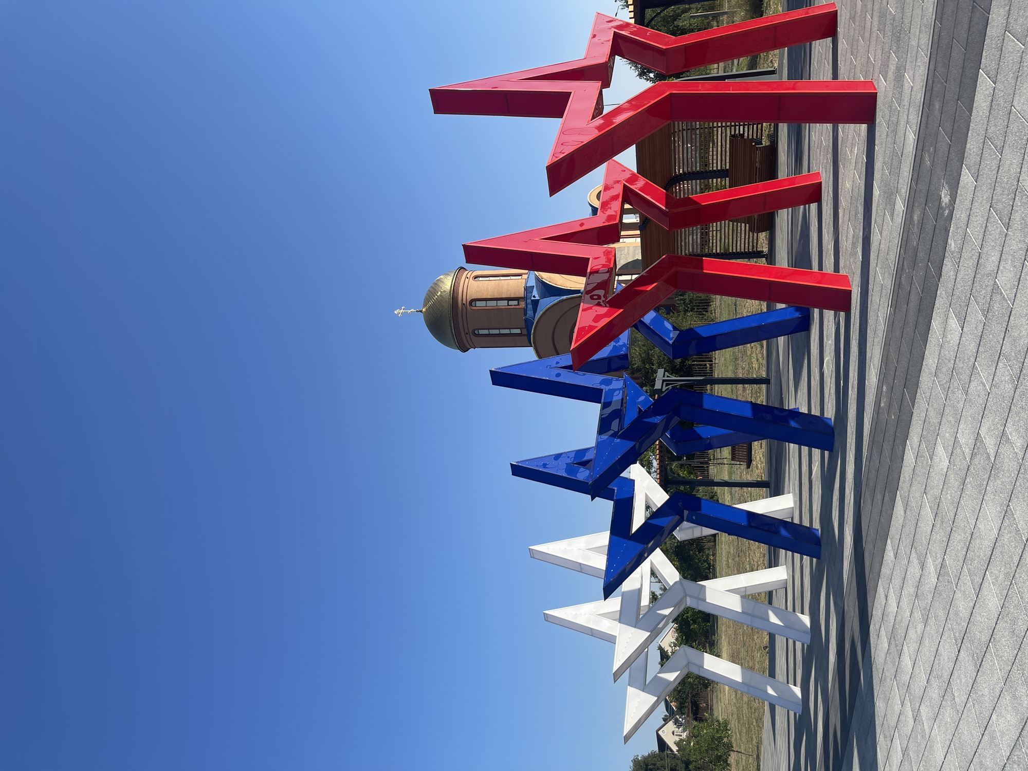 A low-angle, full shot captures a contemporary public art installation of five large, stylized stars in red, white, and blue, set against a clear blue sky. The stars are arranged in a row, with two white stars on the left, followed by two blue stars, and finally two red stars on the right. Behind the art installation, a domed building with a golden cross, resembling a church or Orthodox temple, is visible. The ground in front of the stars is paved with light gray rectangular tiles, and shadows cast by the stars stretch across the pavement. In the background, a wooden structure with a bench can be seen, suggesting a rest area. The surrounding environment includes sparse green grass, trees, and residential buildings to the far left, indicating a semi-urban or park-like setting. The image is taken outdoors during daylight.