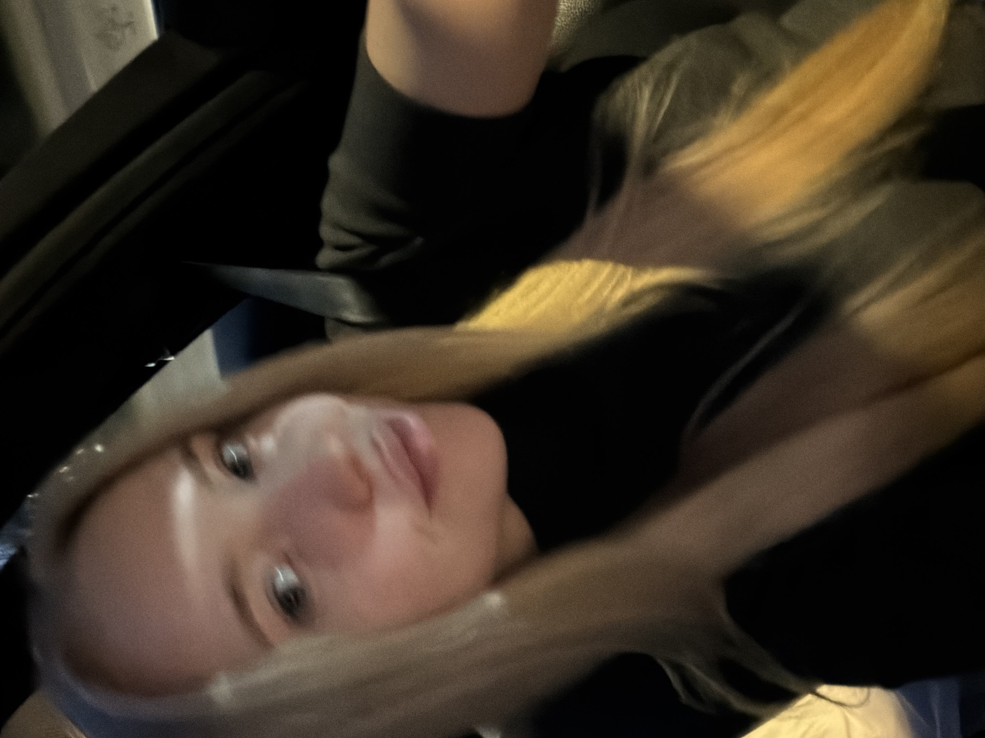 A blurry image depicts a person with blonde hair, light skin, and eyes visible. The person is wearing dark clothing. The background appears to be the interior of a car, with parts of the car door visible on the left side of the frame. The location is unknown.