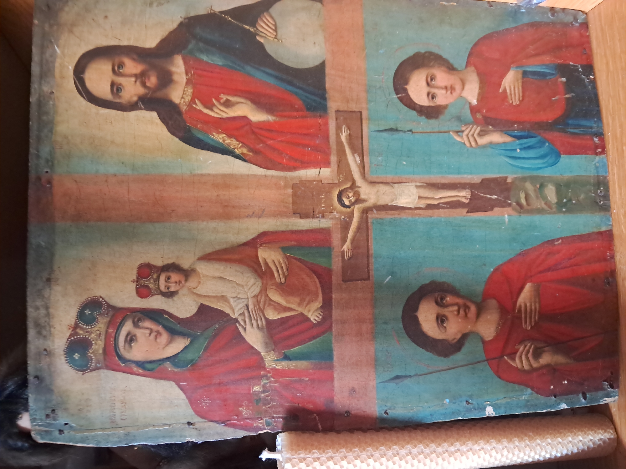 An antique icon on a wooden panel depicts a crucifixion scene. In the upper left quadrant, the Virgin Mary, wearing a crown and red robe, holds the infant Jesus, also crowned. To the upper right, Jesus Christ is shown with a halo, wearing a red robe and blue mantle, with his right hand raised in a blessing gesture. The central part of the icon features a smaller crucifixion scene, with Jesus on the cross. In the lower quadrants, two figures, likely saints, are depicted in red robes, each holding a spear. One of the figures has a halo. The icon is mounted on a wooden panel and appears to be aged, with visible cracks and wear. A stack of beeswax candles is placed to the left of the icon. The icon is located in Smila, Ukraine.