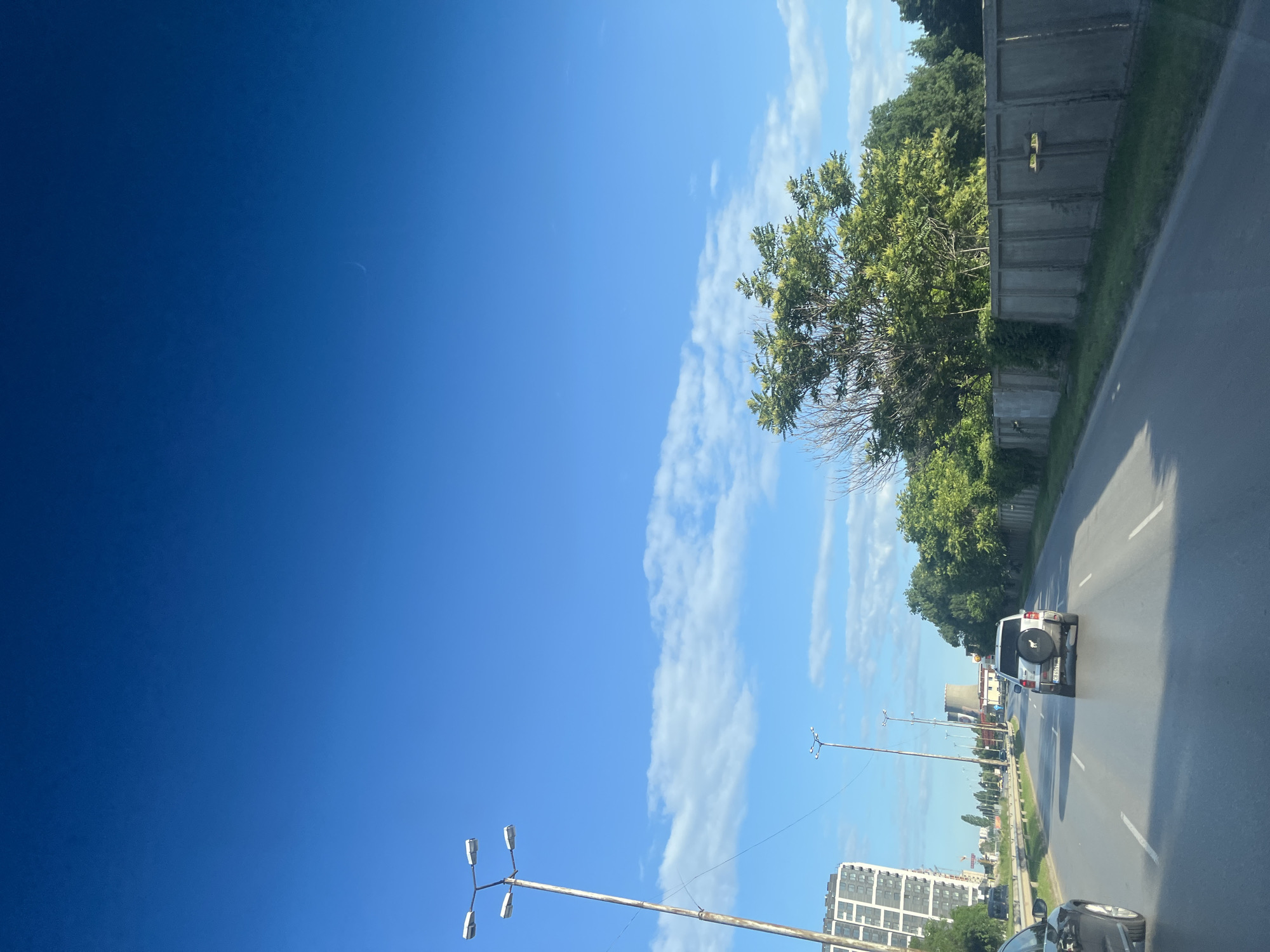 The image shows a clear blue sky with some wispy clouds. In the foreground, a road is visible with a white SUV driving in the right lane. To the left of the road, there is a tall modern building. Further down the road, a cooling tower and other industrial structures can be seen. Lush green trees line the right side of the road, behind a concrete wall. Streetlights are also present along the road. The scene appears to be a suburban or industrial area on a sunny day.