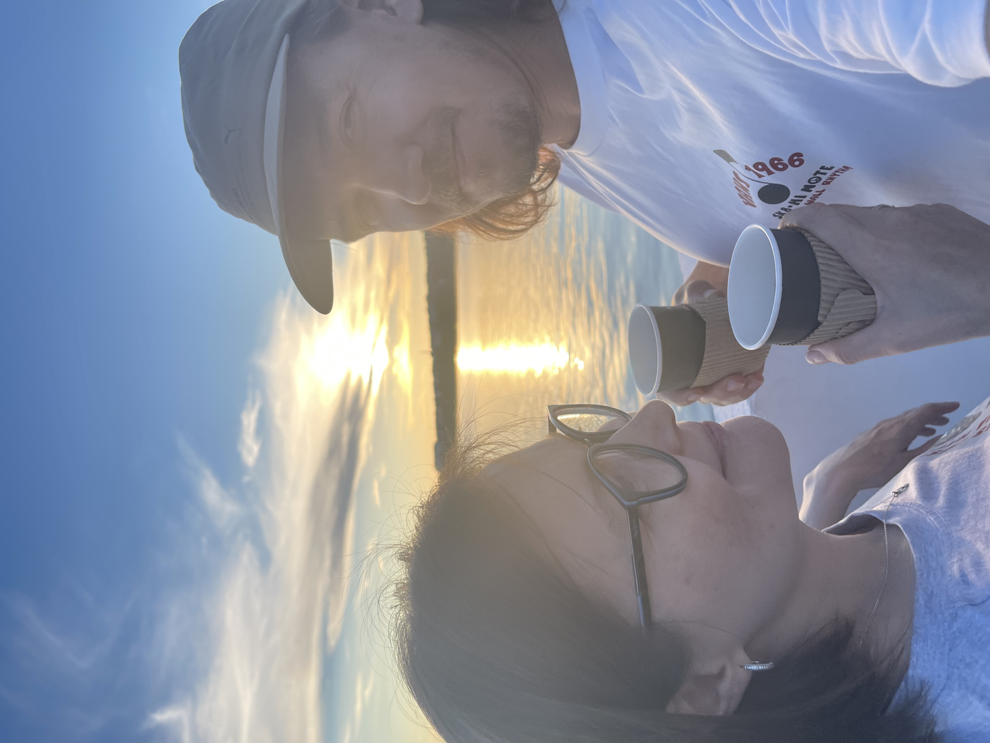 A selfie captures two individuals in the foreground, with a sunset over a body of water in the background. The person on the left, facing right, wears glasses and a gray t-shirt. They are holding a paper cup in their right hand. The person on the right, facing left, wears a light-colored baseball cap and a white t-shirt with a graphic design. They are holding a paper cup in their left hand. The sky is blue with wispy clouds, and the sun reflects brightly on the water. The environment appears to be outdoors, possibly on a boat or by a waterfront, given the water and sky. The overall mood is relaxed, suggesting a shared moment during sunset.