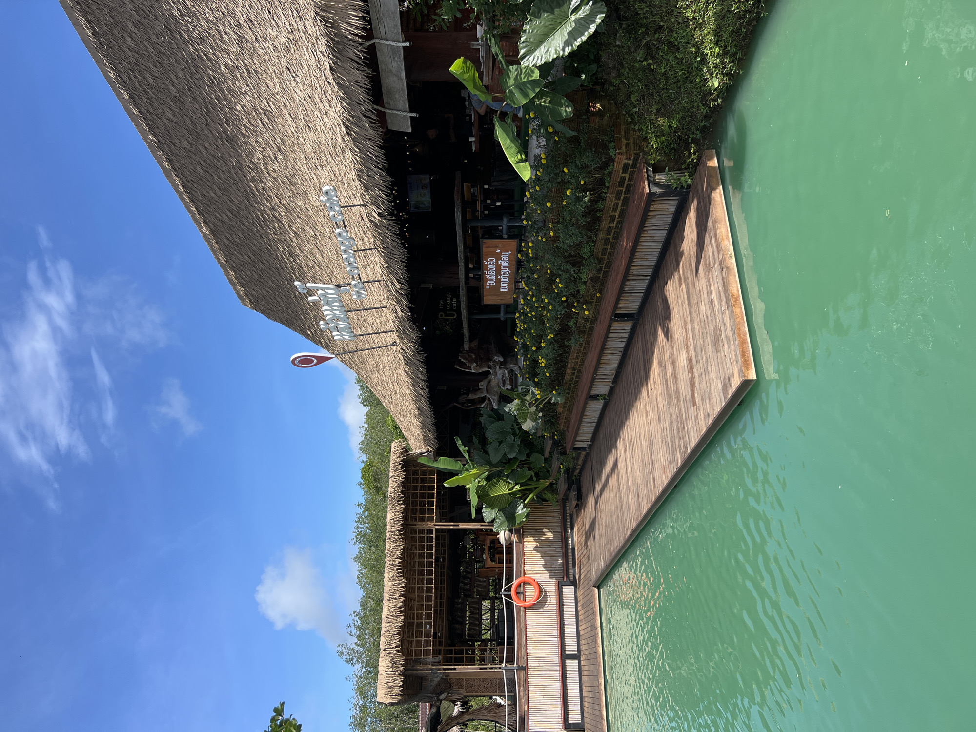 The image depicts a serene waterside cafe with a thatched roof, located in Tambon Pa Kae Bo Hin, Thailand. The cafe features a rustic design with bamboo accents and a wooden deck extending over turquoise water. Lush greenery surrounds the establishment, with a variety of plants and flowers adding to the tropical ambiance. A life preserver is visible hanging on a bamboo post, suggesting proximity to the water. In the background, a dense forest creates a natural backdrop. The sky is a vibrant blue with scattered white clouds, indicating a clear day. A sign with Thai script is displayed on the cafe, likely indicating its name or services. The overall scene conveys a tranquil and inviting atmosphere.