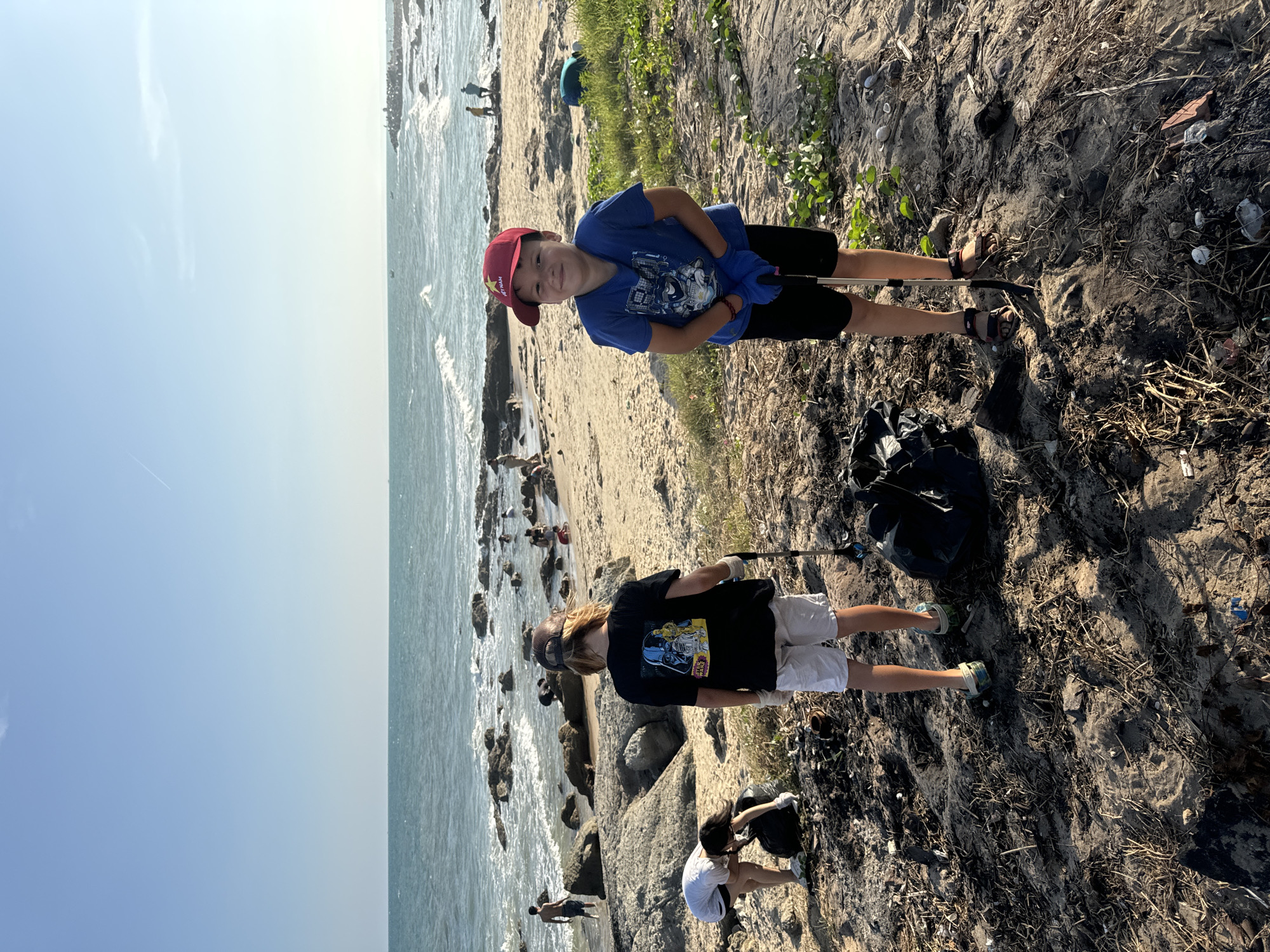 Two children, one wearing a red baseball cap and a blue shirt with black shorts and sandals, and the other with long hair, a baseball cap, black Star Wars t-shirt, and white shorts and athletic shoes, are standing near a black plastic trash bag on a beach in Mũi Né, Vietnam. Both are holding trash-collecting sticks. A third person in a white t-shirt is crouched down near the black trash bag, collecting trash. In the background, people are walking along the beach and wading in the ocean. The ocean is visible on the left side of the image, with a blue sky above. The beach has a mix of sand and seaweed.