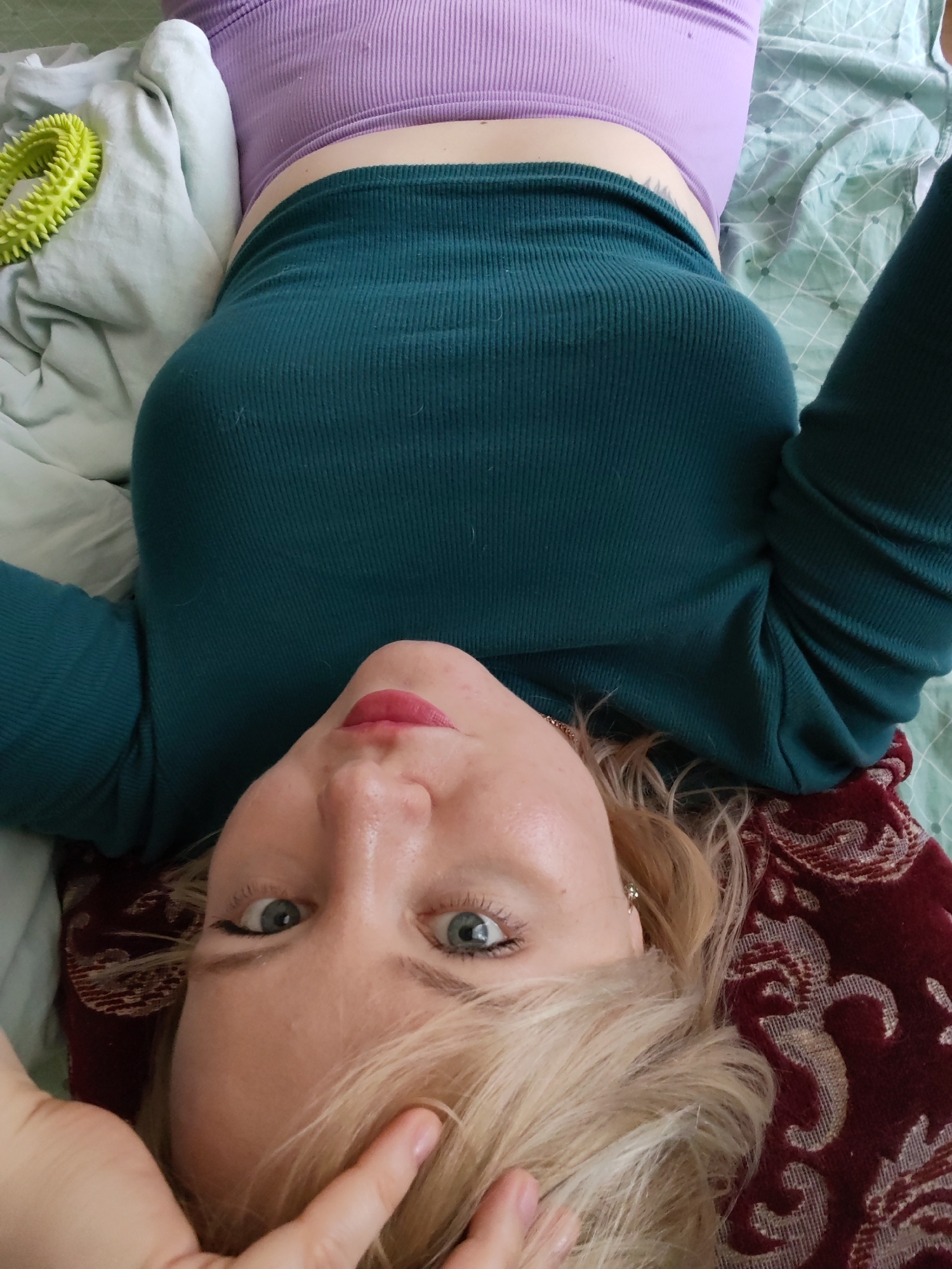 This overhead photograph captures a woman with blonde hair and blue eyes, lying on her back on a bed, looking directly at the camera. She has a fair complexion, subtly made-up eyes with eyeliner, and lips with a pinkish tint, displaying a calm and relaxed expression. She is dressed in a dark teal, long-sleeved, ribbed top, under which a light purple ribbed crop top is visible, exposing a portion of her midriff. Her lower body is covered by what appears to be matching dark teal ribbed pants or leggings. Her right hand is visible near her head, lightly touching her blonde hair.
The setting is an indoor space, likely a bedroom, with the woman positioned on a bed. The bedding consists of a light blue-green sheet with a subtle checkered pattern and a crumpled light-colored blanket or sheet. To her left, a bright yellow-green spiky, ring-shaped object is visible on the bed, possibly a massage tool. Her head rests on a dark red pillow adorned with an ornate gold or beige pattern. There are no visual cues to indicate the specific time of day or outdoor weather conditions. The photograph was taken in Izhevsk, Russia. No text is visible within the image.