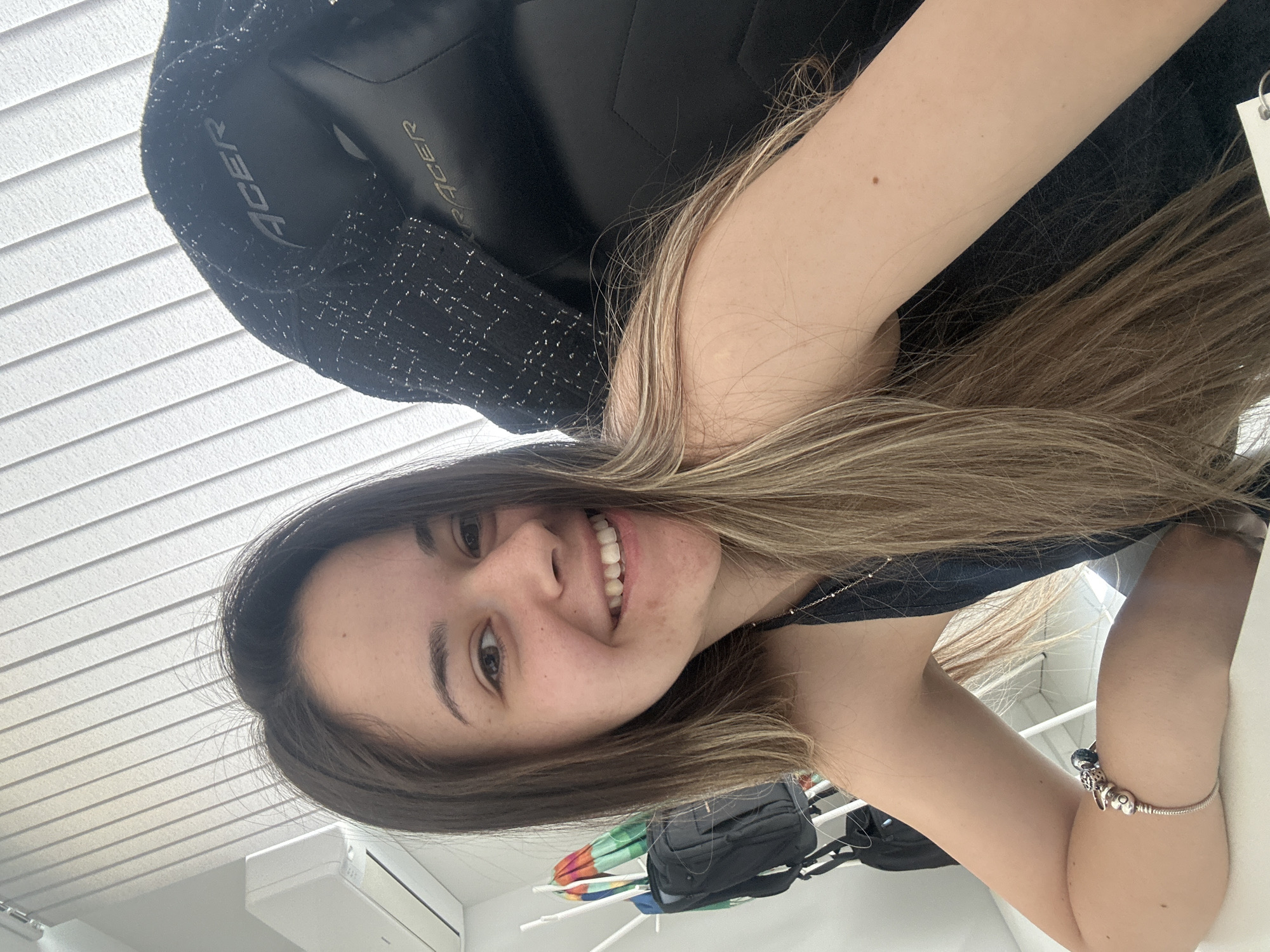 A young woman with long, light brown and blonde hair smiles at the camera. She is sitting in an Acer gaming chair with a black fabric back. She is wearing a black top and a silver bracelet is visible on her left wrist. The background consists of white horizontal blinds and what appears to be part of a room with a white wall, an air conditioning unit, and possibly a backpack and a laundry rack with some colorful items. The photo does not provide location information for Sofia, Bulgaria.