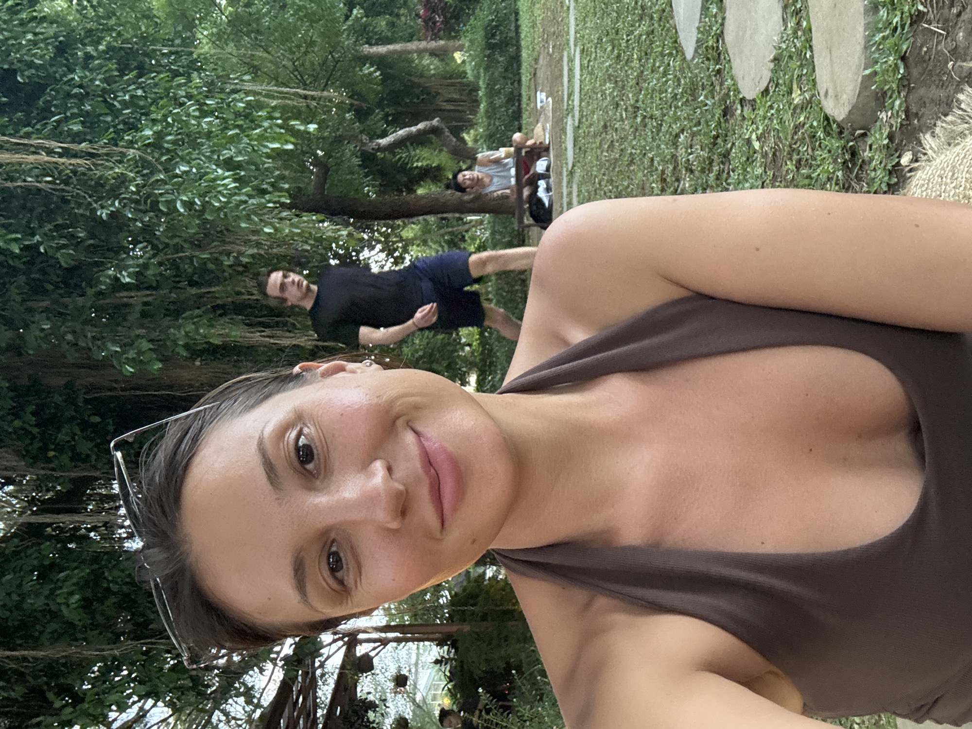 The image is a selfie taken outdoors, likely in a park or garden setting. The foreground is dominated by a woman with brown hair and sunglasses perched on her head. She is wearing a brown halter top.
In the background, two men are visible. One is walking, wearing a black shirt and blue shorts. The other is lying down on a mat.
The environment includes trees, grass, and stone or concrete slabs arranged in a pathway. The location is Pecatu, Indonesia.