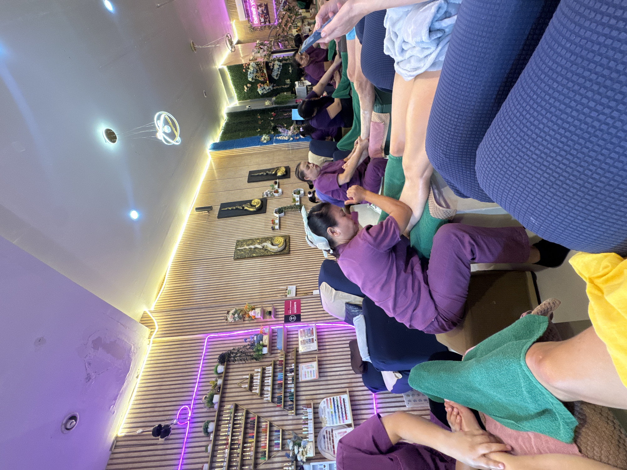 The image shows the interior of a business, likely a spa or massage parlor, located in Patong, Thailand. Several people, likely customers and employees, are visible. Employees wearing purple uniforms are attending to customers who are seated in cushioned chairs. Some customers are receiving foot massages, while others appear to be engaged in activities such as using mobile phones. The environment is decorated with wooden panels, artificial plants, and LED lighting. Shelves displaying various products, possibly nail polishes or lotions, are also visible.