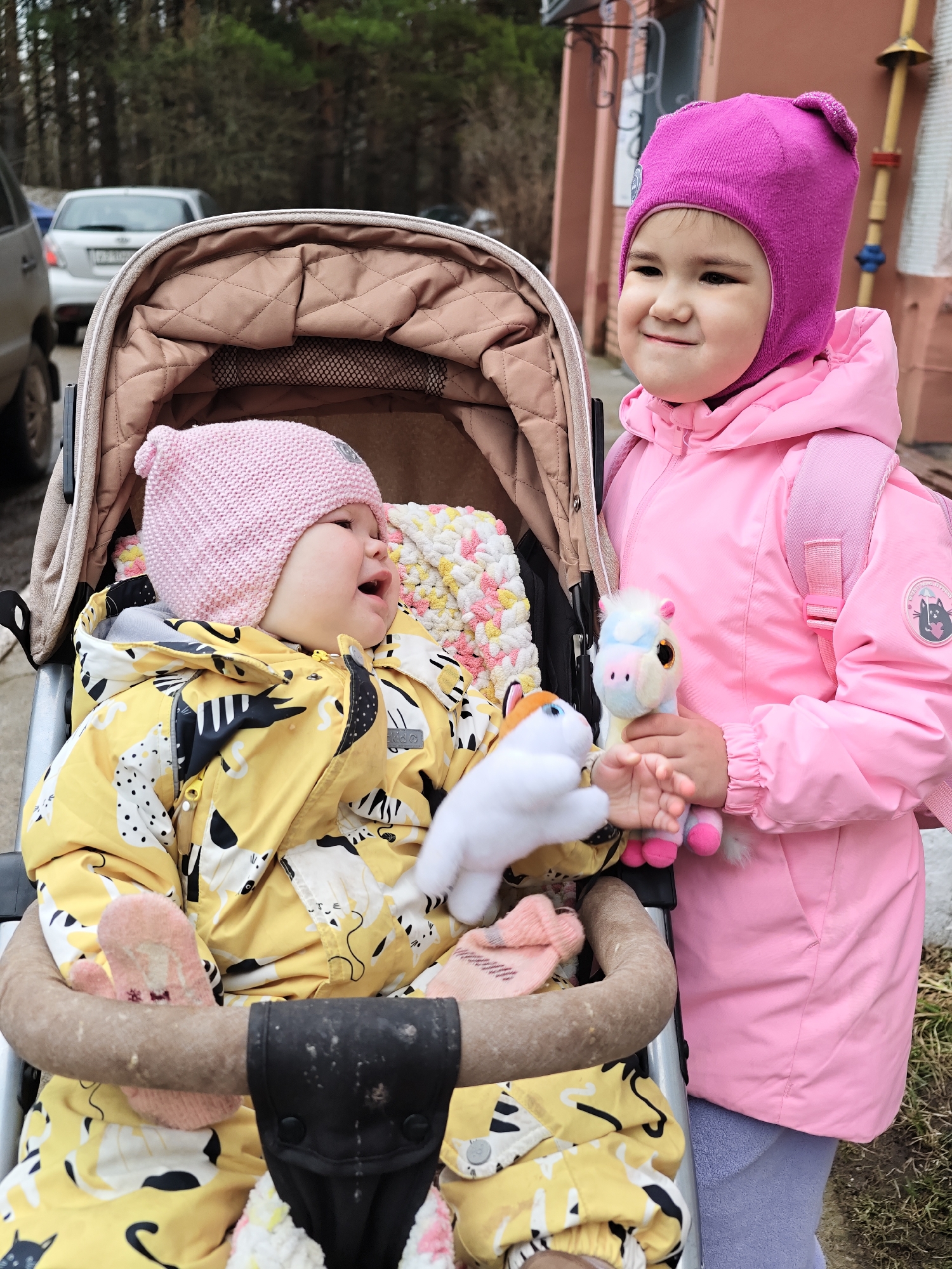 Two young girls are outdoors near what appears to be a building and some parked cars. One girl, wearing a pink hat and pink jacket with a small backpack, is standing next to a stroller. She is holding two small stuffed animals. The other girl is sitting in the stroller. She is wearing a pink hat and a yellow jacket with a pattern of cats. Her mouth is open, as if she is talking or crying. The stroller has a tan-colored interior. The background shows cars and a building. The location is identified as Chaikovsky, Russia.