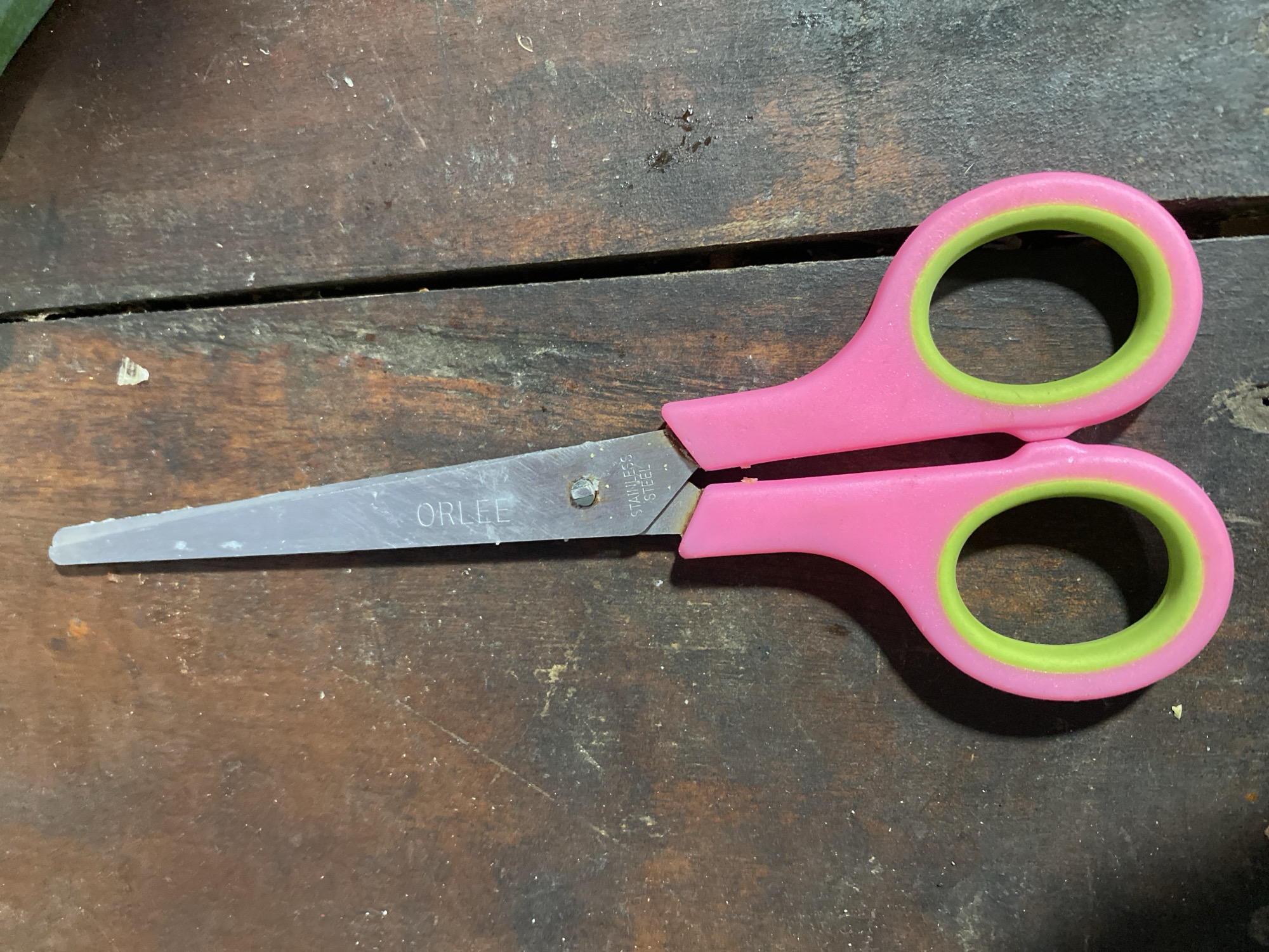 A pair of pink-handled scissors with green grips lies open on a weathered wooden table. The scissors' metal blades are visible, with the word "ORLEE" imprinted on one blade and "STAINLESS STEEL" on the other. The table's surface is rough, marked with visible grain and some debris. The lighting suggests an indoor setting. The location is a domestic or workshop environment within the city of Kasihan, Indonesia.