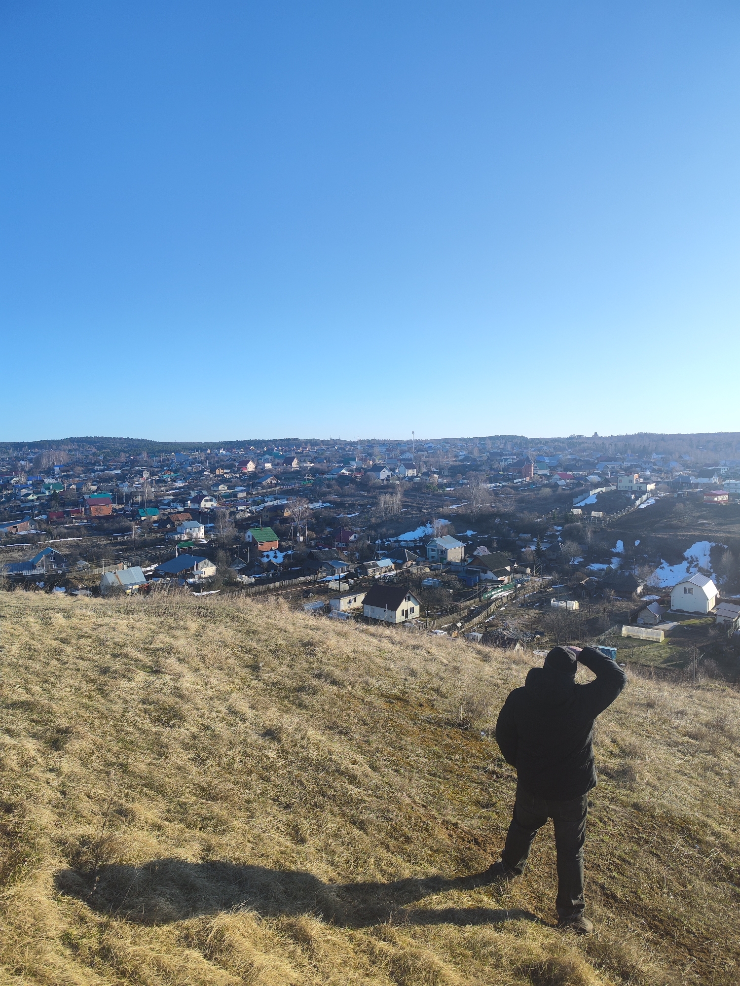 A man stands on a grassy hillside, overlooking a dense urban area in Noviy, Russia. The man is wearing dark pants and a dark jacket, with his right hand shielding his eyes as he looks towards the cityscape. The terrain beneath him consists of dry, brown grass. The city below is composed of numerous residential buildings of varying sizes and colors. The sky above is clear and blue.