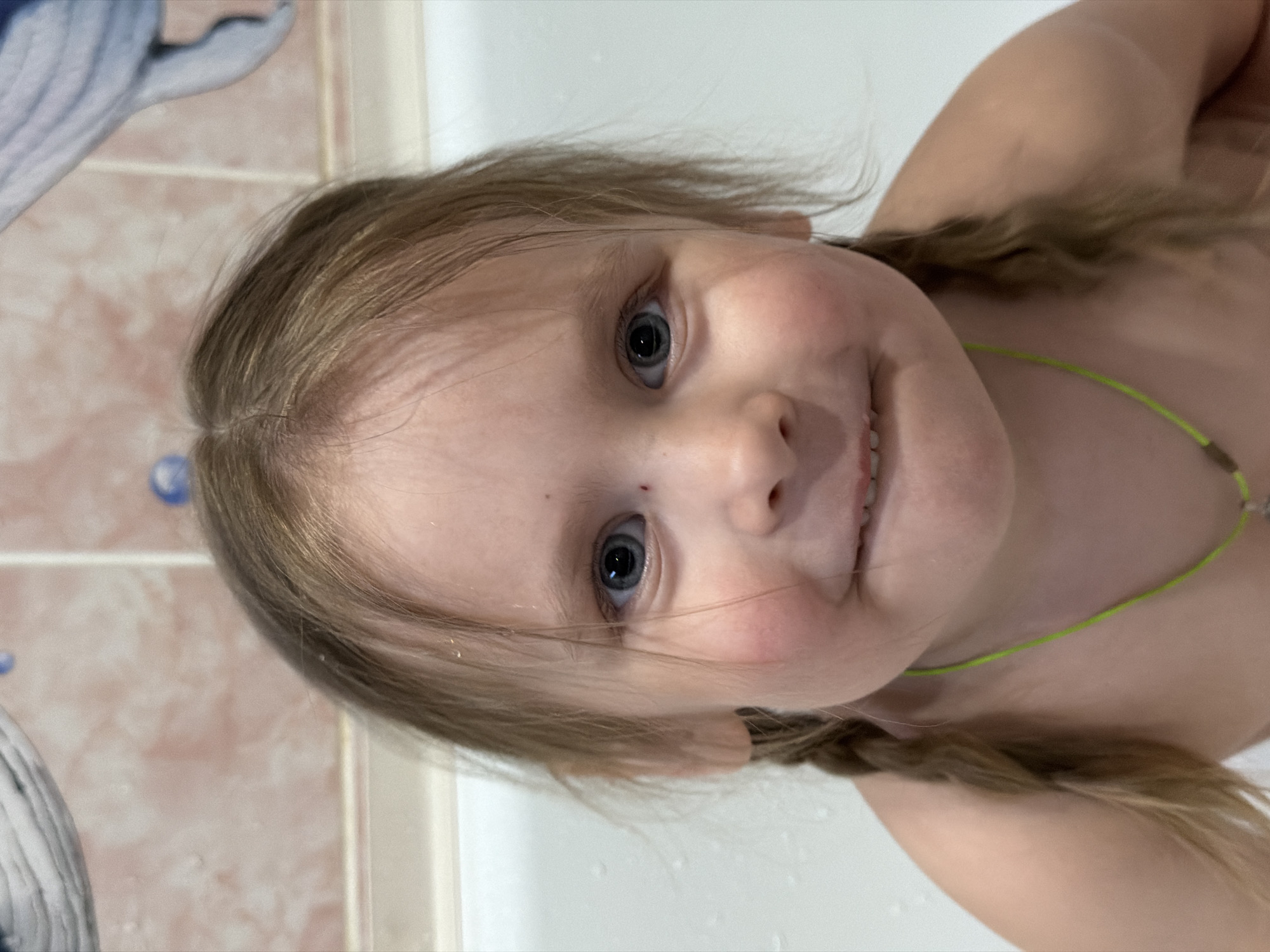 A young child with light skin and light brown hair braided into pigtails is photographed from a slightly high angle, turned on their side. The child has light eyes, and a small mark is visible on their forehead. They appear to be in a bathtub or shower setting, with white surfaces visible. A bright green cord necklace is around their neck. The background shows tiled walls with a faded pattern, and a white and blue object, possibly a toy, is partially visible in the upper left corner. The location in the city of Chaikovsky, Russia is not evident from the image itself.