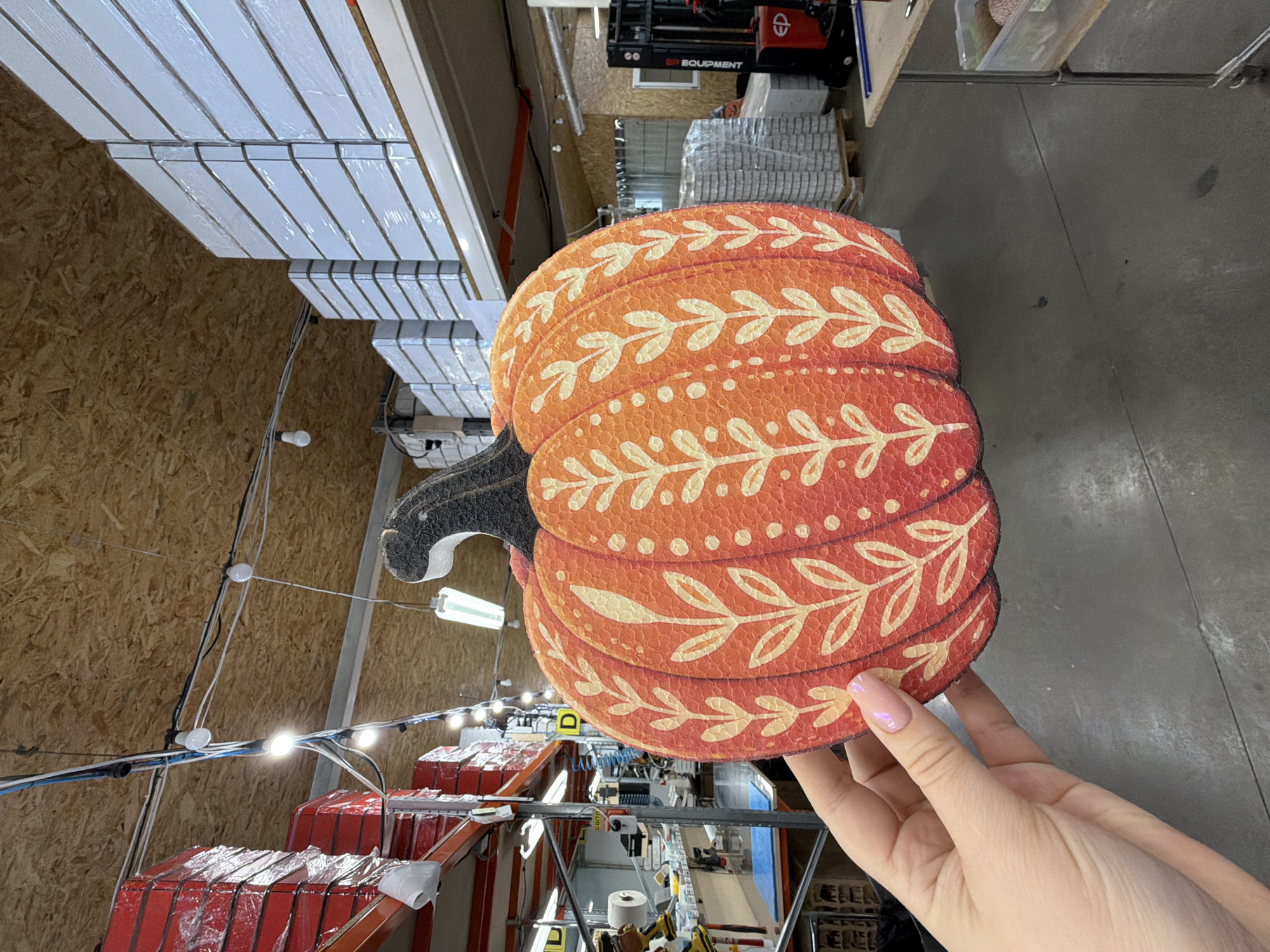 A person's hand, with a pink, iridescent nail polish, holds up a decorative, orange, pumpkin-shaped cutout. The cutout is adorned with a repeating pattern of stylized, white leafy branches and small white dots. The background suggests an indoor storage or warehouse environment, with tall shelves filled with stacked white boxes on the right and red-wrapped items on shelves to the left. Overhead, exposed wooden ceiling beams are visible, along with a network of electrical wires and lights. A forklift is present in the middle ground to the right. The floor appears to be concrete.