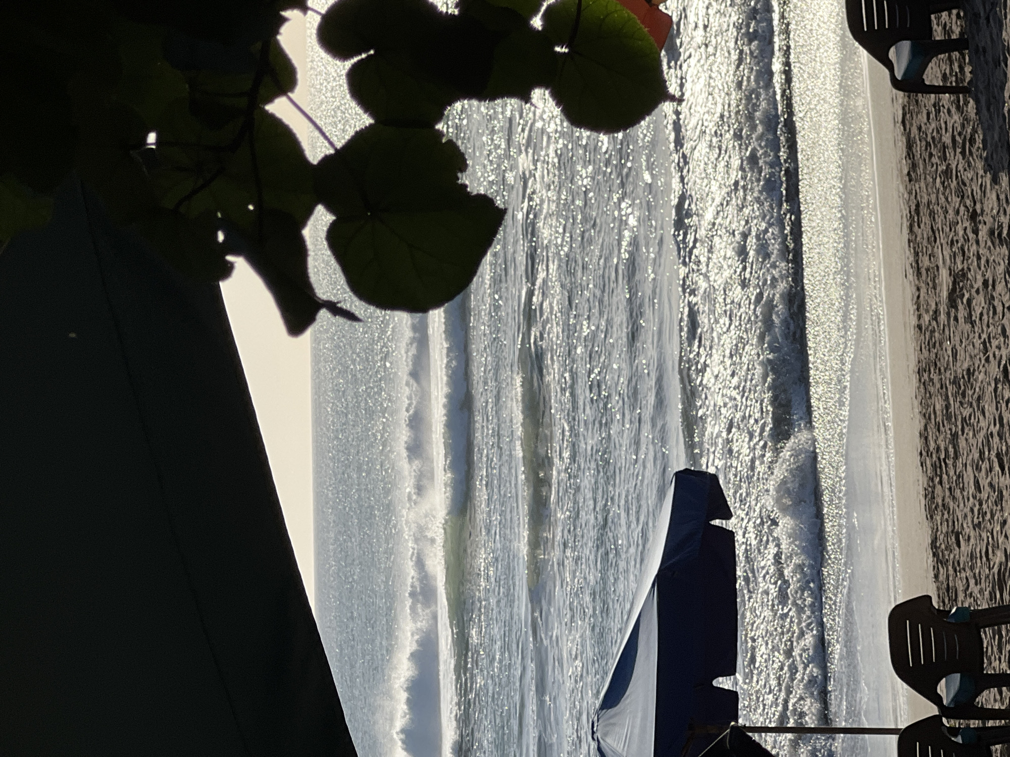 The image depicts a beach scene at Kuta, Indonesia. The foreground shows a sandy beach with two dark plastic chairs and a portion of a dark blue beach umbrella. In the middle ground, the ocean is visible with several waves breaking near the shore, reflecting the bright sunlight. The background is the ocean meeting the horizon under a bright sky. Silhouetted leaves and branches of a plant hang from the upper right corner, and a dark structure, possibly another umbrella or awning, juts in from the upper left. The overall lighting suggests it is daytime, likely morning or late afternoon due to the angle of the sun and the shadows. There are no people visible in the frame.