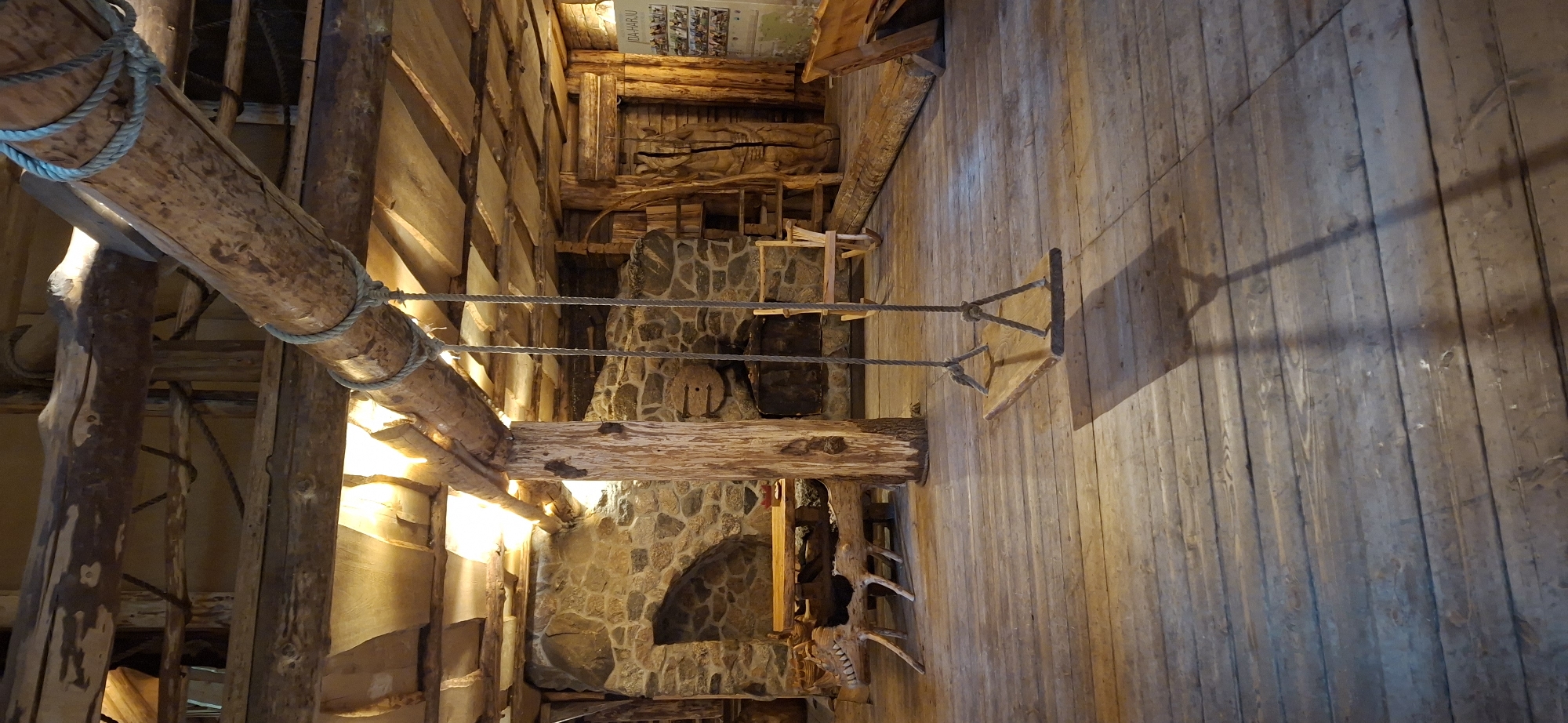 The image shows the interior of a rustic wooden building, likely a lodge or a recreational facility, located in Saula, Estonia. A wooden swing hangs from a large log beam suspended across the space. The room is constructed primarily of wood, with rough-hewn logs forming structural elements and a wooden plank floor. Stone walls are visible in the background, suggesting a fireplace or an older structure integrated into the design. Various wooden furniture pieces, including a table and benches, are scattered throughout the space. The lighting appears to be from overhead sources, casting warm light and shadows. The overall atmosphere is cozy and natural, emphasizing the use of raw, natural materials. No people are present in the image.