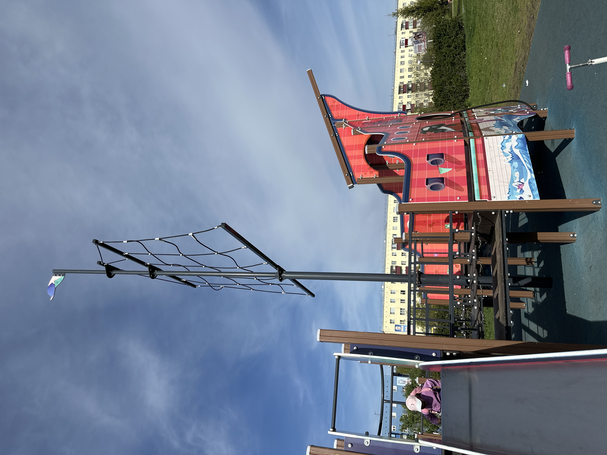 Here is a description of the image:
The image captures a playground setting with a ship-themed structure as the main element. The structure is predominantly red with brown wooden supports and includes detailing such as porthole cutouts and marine-themed graphics. Adjacent to the ship is a climbing net supported by a tall metal pole with a small flag attached. A slide is partially visible in the foreground, with a person wearing a purple jacket and a pink cap at the top, seemingly preparing to slide down.
In the background, a building with multiple windows is seen, suggesting an urban or residential area. A patch of green grass further defines the setting. The sky above is blue with light cloud cover.