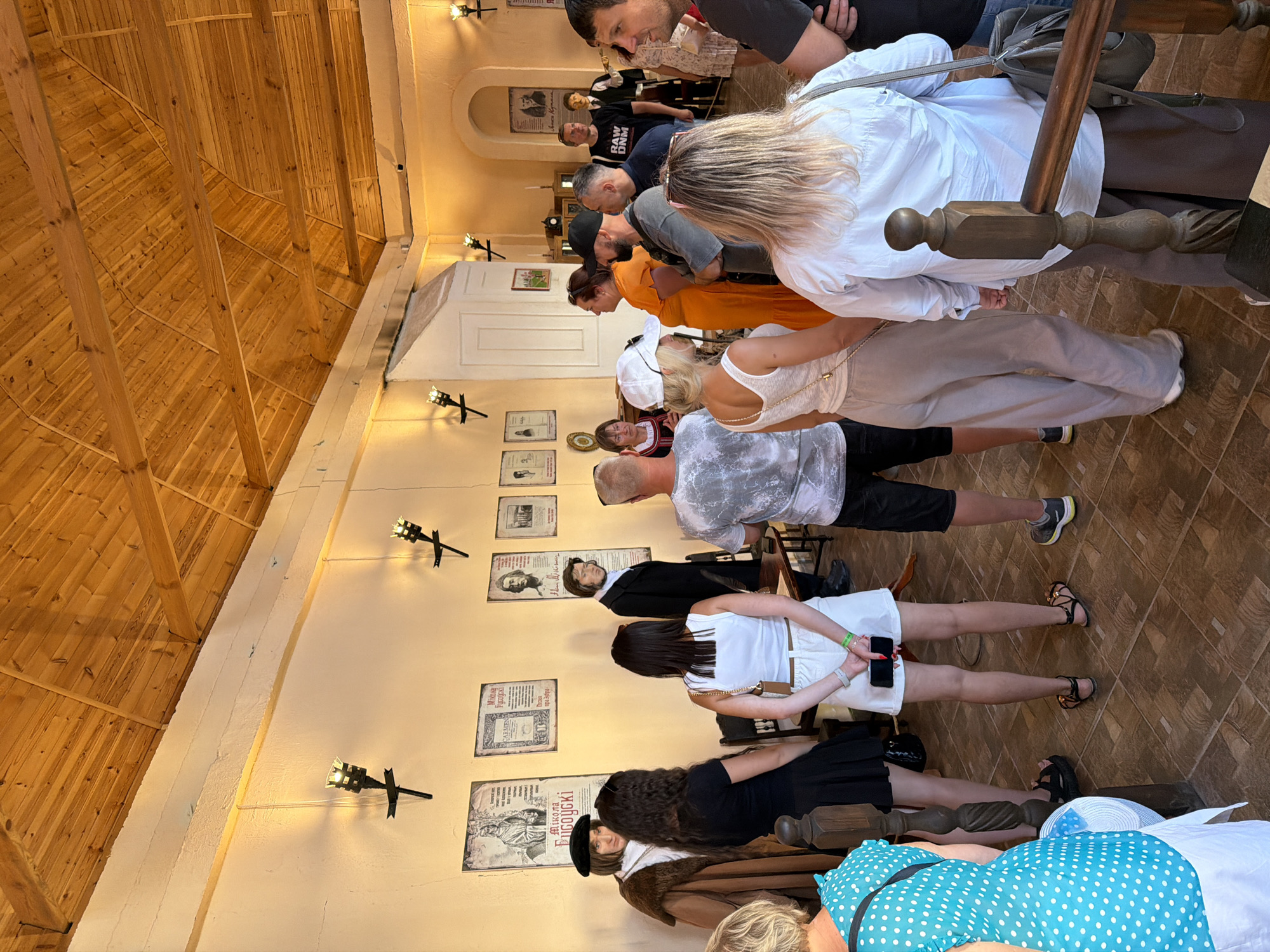 The image depicts an indoor gathering of approximately 15 individuals, appearing to be tourists, in a room with a wooden slatted ceiling and light-colored walls adorned with framed documents and wall sconces. Several mannequins are positioned around the room, dressed in historical attire, suggesting the location might be a museum or historical exhibit. Some individuals are standing and observing the exhibits, while others are engaged in conversation. The flooring is tiled in a brown, diamond pattern. The overall atmosphere suggests a visit to a cultural or historical site.