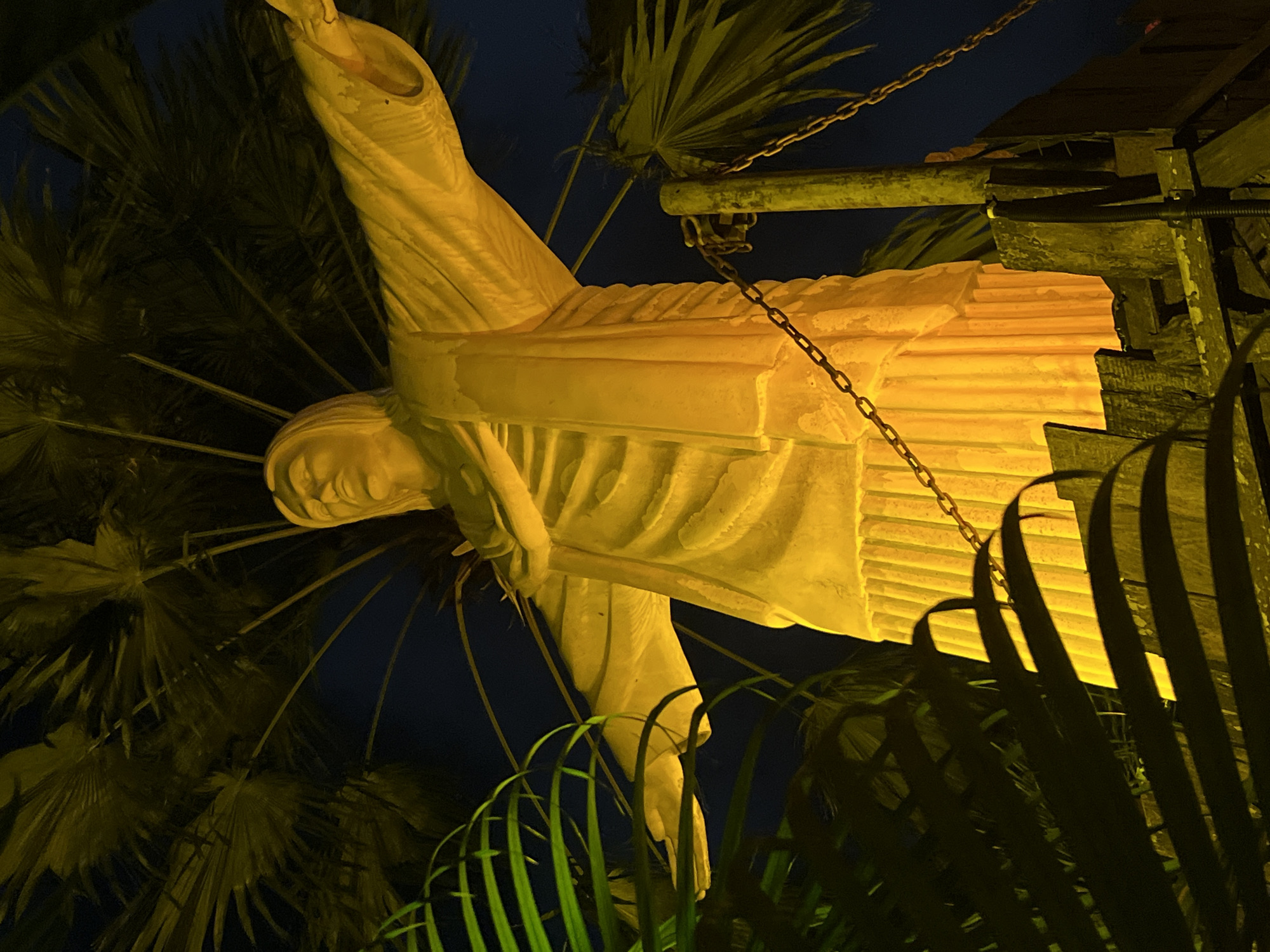 A low-angle, full-body shot depicts a statue of Christ the Redeemer, illuminated by a golden light, set against a dark sky and surrounded by tropical foliage, likely palm fronds. The statue is positioned on a raised wooden platform, with visible chains attached to a wooden post to its right. The statue's arms are outstretched in a characteristic pose, and its robe is rendered with detailed folds. The image appears to be taken at night, with the artificial lighting highlighting the statue and the surrounding vegetation. The presence of the statue and tropical plants suggests a location in a warm climate, potentially Bali, Indonesia, given the request to include Kuta.