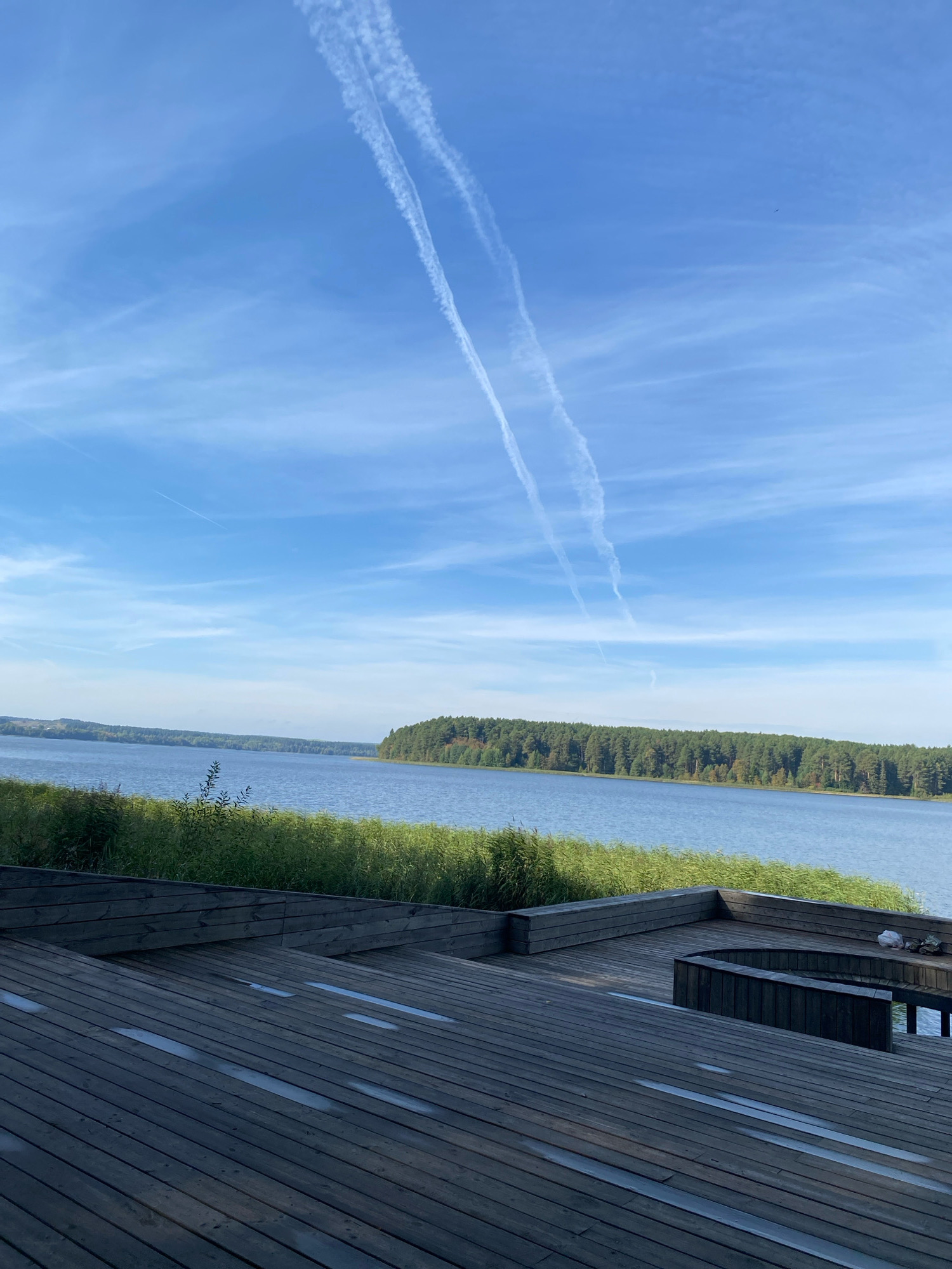 The image depicts a serene outdoor scene with a focus on a body of water and a wooden deck. In the foreground, a weathered wooden deck with visible planking and some damp patches extends diagonally across the frame. The deck appears to be tiered, with some built-in wooden seating structures visible. Behind the deck, a dense line of green reeds or tall grass forms a natural barrier.
Beyond the reeds, a calm, expansive body of water stretches towards a distant, tree-covered shoreline. The trees appear to be a mix of coniferous and deciduous, with shades of green and hints of autumnal colors. The water is a deep blue, reflecting the sky above.
The sky is a clear, bright blue with scattered, wispy white clouds and two prominent contrails from an aircraft streaking across the upper portion of the image. The lighting suggests it is daytime, likely mid-morning or afternoon.
The scene appears to be a public or private waterfront area, possibly a recreational spot. There are no people or animals prominently visible in the image, though a small white object, possibly a piece of litter or a stray item, is present on the wooden structure near the right edge. The location is not explicitly identifiable as Votkinsk, Russia, based solely on the visual information provided.