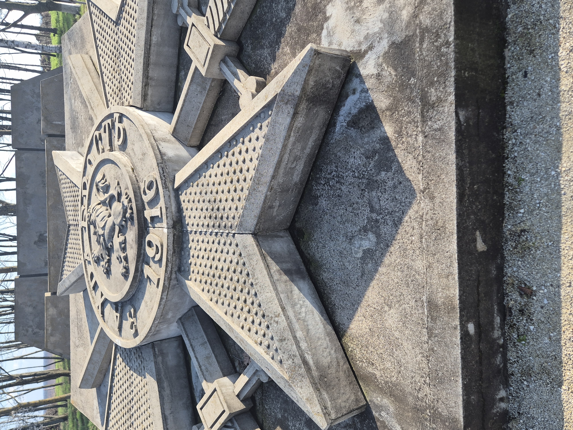 The image shows a stone monument in Shumentsi, Bulgaria. The monument is a large, stylized Maltese cross with a circular medallion in the center. The medallion features a relief image and the inscription "1916." The cross has a textured surface with small, rounded protrusions. The monument is made of grey stone and is partially in shadow. The background includes trees and a gravel surface.