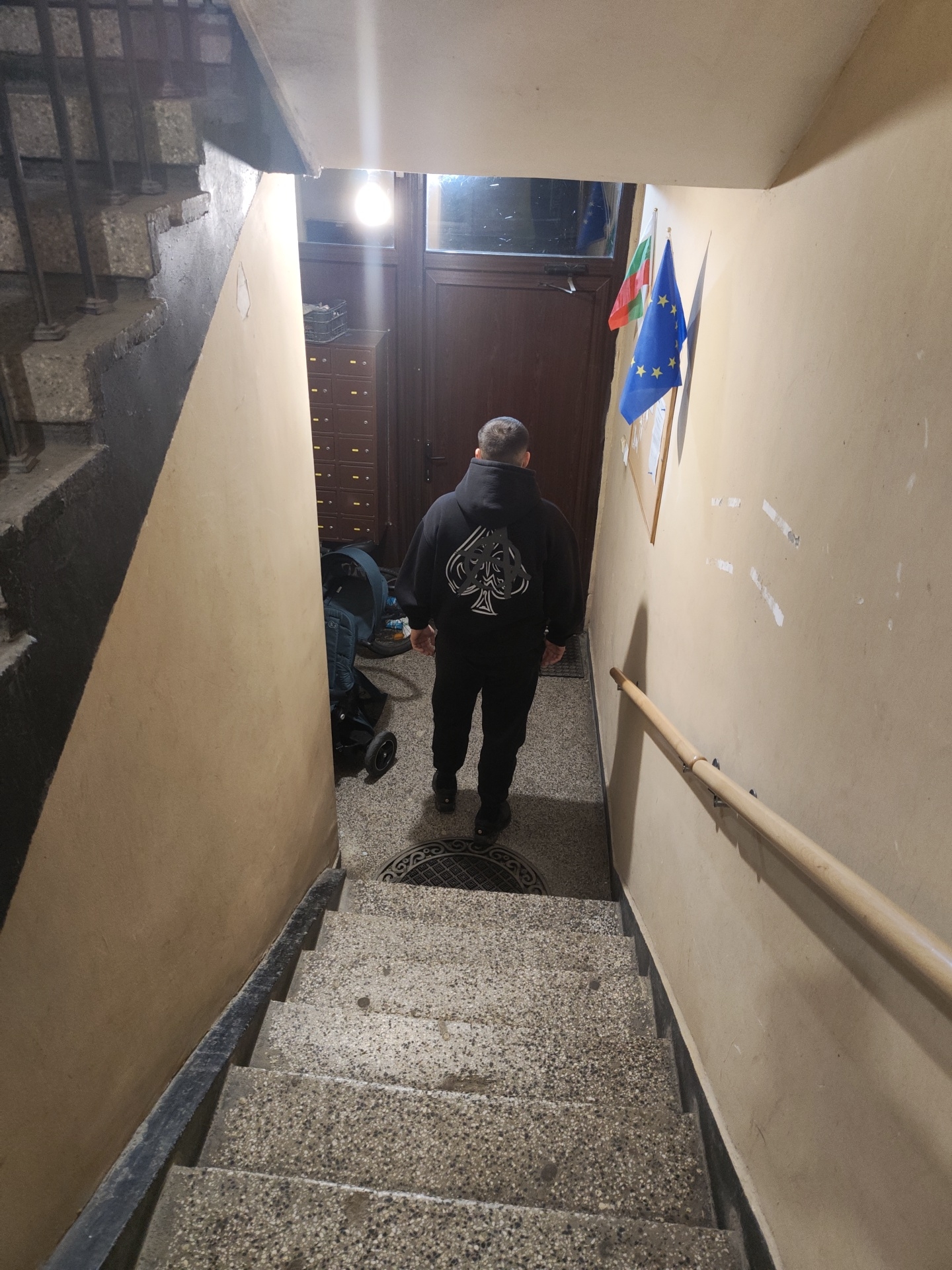 A medium shot captures a man standing at the top of a flight of concrete stairs, appearing to look down. He is dressed in a black hoodie with a white spade symbol on the back and black pants. To his left, a baby stroller is parked near a wall of mailboxes. Behind him, through a doorway, there is a bright light. To his right, the wall has two flags hanging, one of Bulgaria and one of the European Union. A wooden handrail is mounted on the wall along the stairs. The stairs themselves are made of speckled concrete, and the walls are painted a faded yellow color. Based on the details of flags hanging and the design of the building, this scene is likely in Kozloduy, Bulgaria.