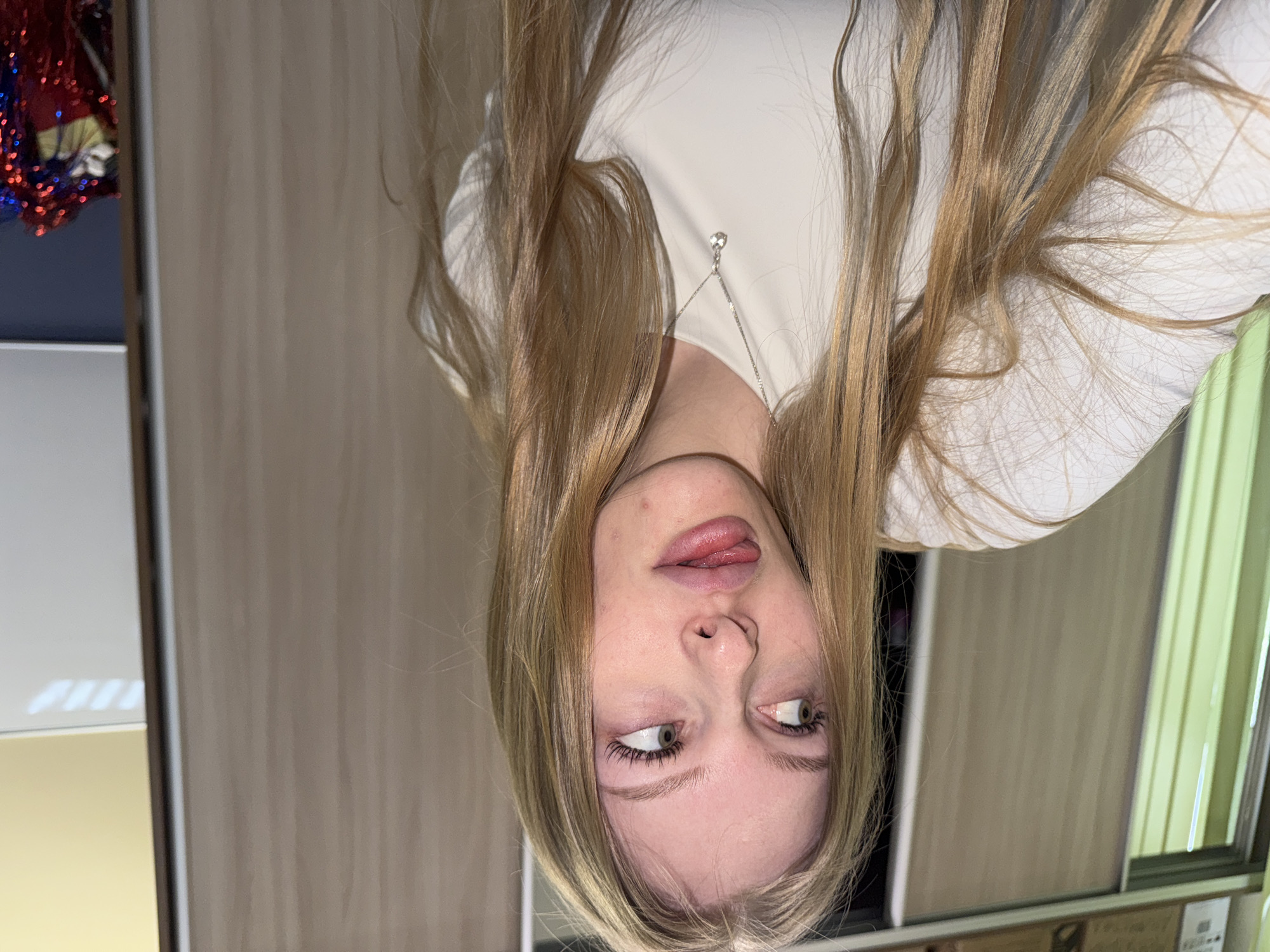 A young woman with long blonde hair is upside down in the frame. She wears a white top and a silver necklace. She has her tongue sticking out and her eyes are rolled. The background includes a cabinet or wardrobe with light brown doors and a decorative garland on top of a dark blue surface. No indication to suggest this photo was taken in Perm, Russia.