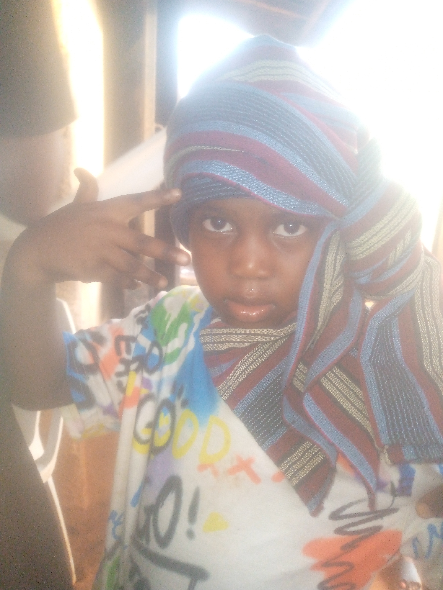 A young child, possibly a boy, with dark skin, is centered in the frame, looking directly at the viewer. The child is wearing a headwrap made of striped fabric in dark blue, maroon, and lighter beige or gold colors. The headwrap covers the child's hair and wraps around their neck. They are also wearing a white t-shirt featuring colorful, graffiti-style text and designs; visible text fragments include "GOOD" and "GO!".
The child's right hand is raised to their head, forming a "finger gun" gesture with the index finger extended and the thumb raised. Their expression is serious or focused, with slightly parted lips. The background is heavily overexposed and very bright, suggesting strong backlighting, possibly from a window or an outdoor light source. Faint vertical structures are barely visible in the bright background. A portion of another person's arm or shoulder can be seen on the far left of the image. The overall bright lighting indicates it is daytime. The specific location is not discernible.