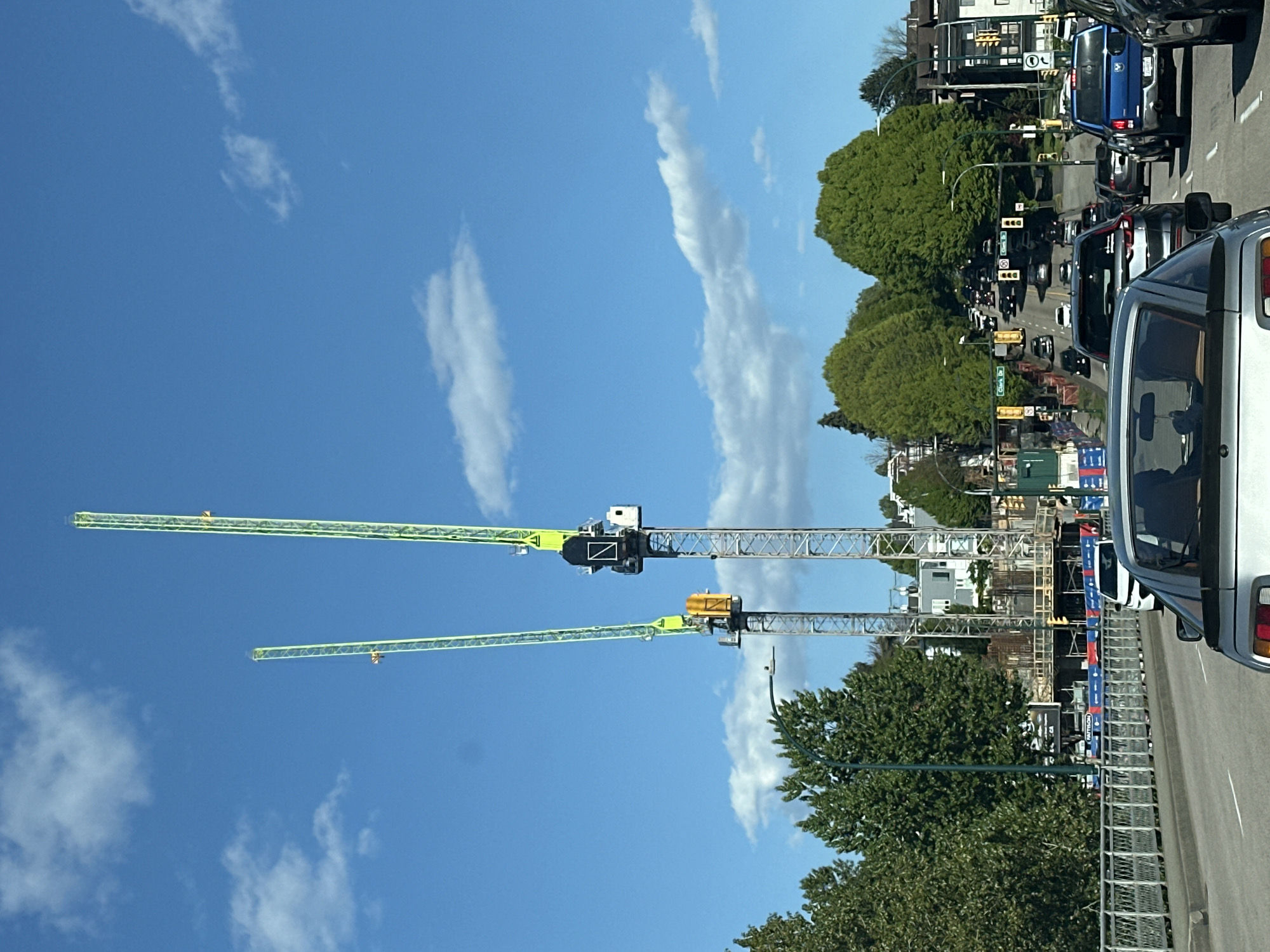 The image depicts a street scene in Vancouver, Canada, marked by the presence of two construction cranes in the foreground. The cranes are characterized by their long, green and silver lattice booms extending across a blue sky with scattered white clouds. Below the cranes, there is a partial view of a construction site with steel frameworks and barriers. A row of trees lines the street, partially obscuring the view of buildings in the background. The right side of the image shows a busy street with multiple cars moving in the same direction. The overall atmosphere is urban, likely during daytime, with active construction activities.