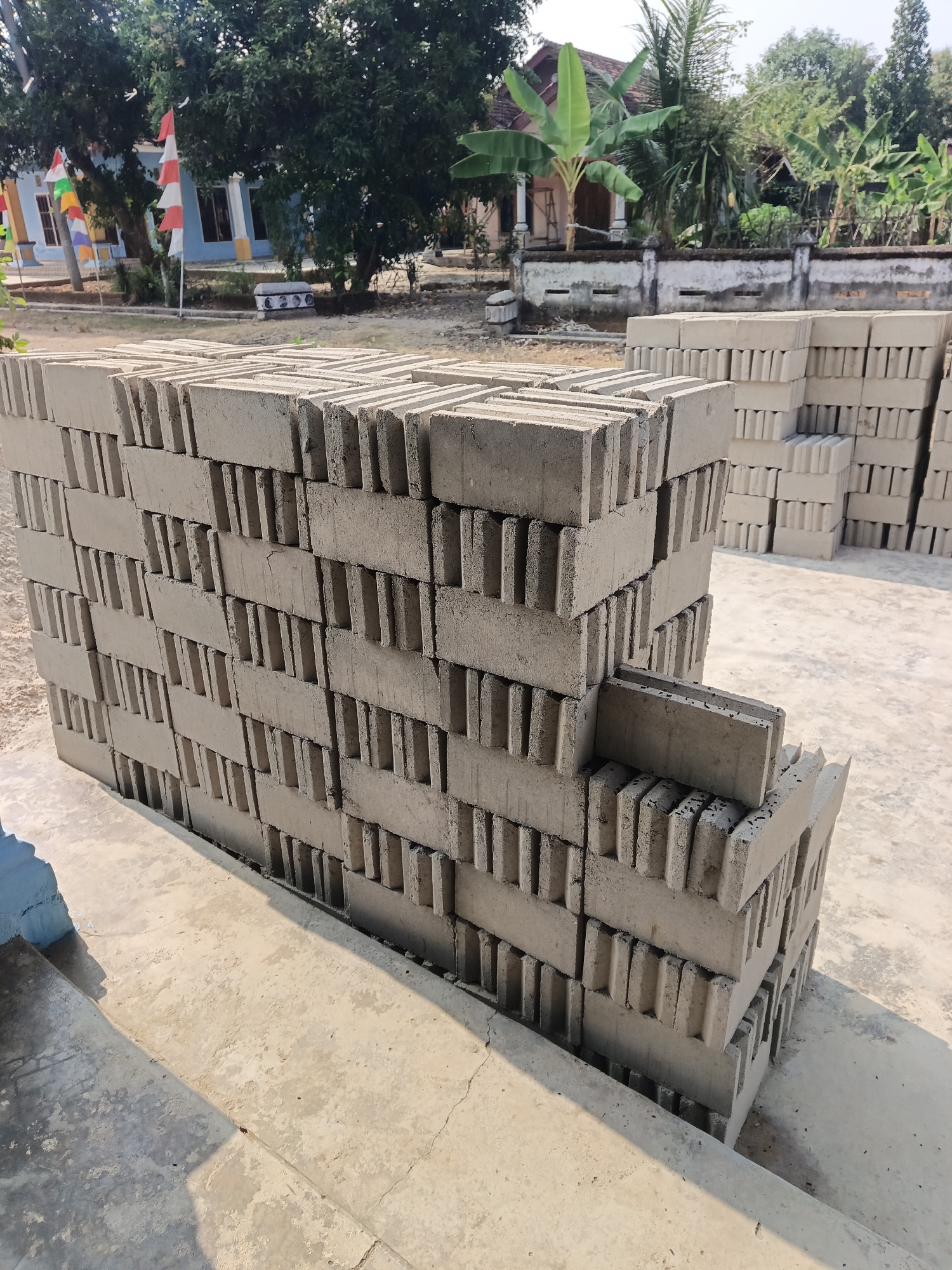 The image shows a close-up view of stacks of concrete blocks on a paved surface. In the background, there is a building with blue walls and a red and white flag. Trees and houses are also visible. The scene appears to be outdoors, possibly in a construction or storage area. There are no people or animals visible in the image. The lighting suggests it is daytime.
