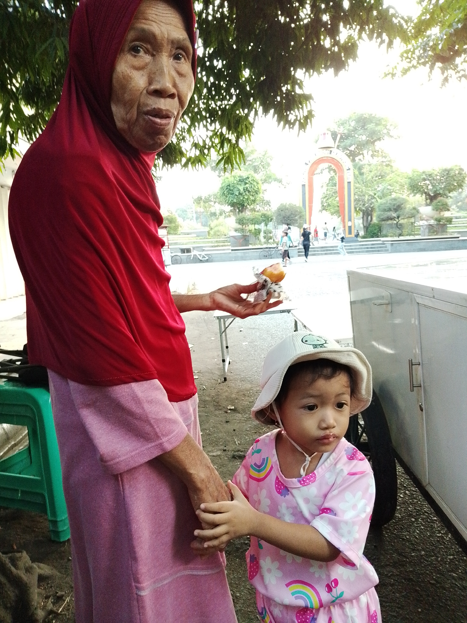 An elderly woman wearing a red hijab and a pink dress is holding a small food item wrapped in plastic. Beside her stands a young child in a pink outfit and a white hat. The woman's right hand is extended, holding the food, while her left hand appears to be touching the child's hand or arm. The child is looking to the right. They are standing on a paved surface, possibly a sidewalk or public area, with a white cart or food stall to their right. In the background, there are trees, a decorative archway, and several people walking. The overall lighting suggests it is daytime, possibly late afternoon. The scene appears to be a public space within Kecamatan Pati, Indonesia, given the architectural style of the archway and the presence of vegetation common in tropical environments.