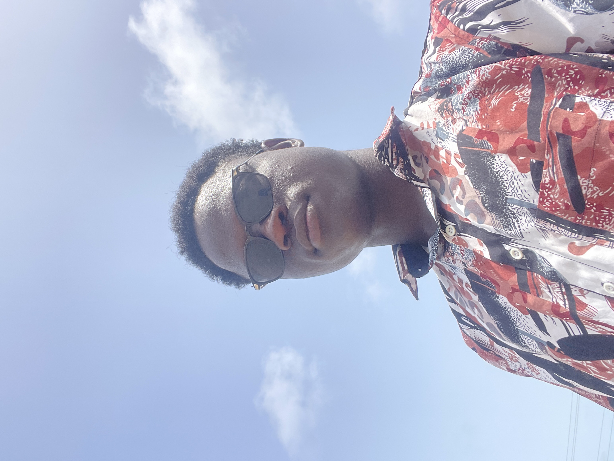 A low-angle shot of a person wearing sunglasses and a patterned button-down shirt against a bright, blue sky with scattered clouds. The person is looking down towards the camera. The setting appears to be outdoors during daylight hours. Specific location details within Badagry, Nigeria, are not discernible from the image.