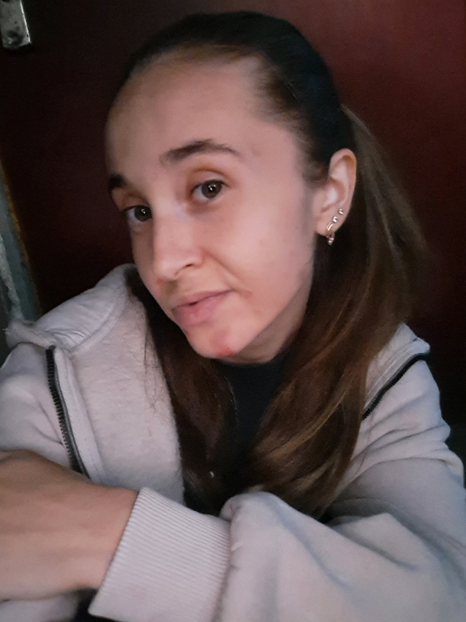 A young woman with light brown hair pulled back in a ponytail and fair skin is captured in a close-up, bust shot. She is wearing a light-colored hooded jacket with a zipper. Her left arm is crossed over her chest. She has two small earrings in her left ear, with the lower one appearing to be a hoop. There are some small red marks on her chin. The background is dark and appears to be a wall. The image does not contain any information about the location within Vypasne, Ukraine.