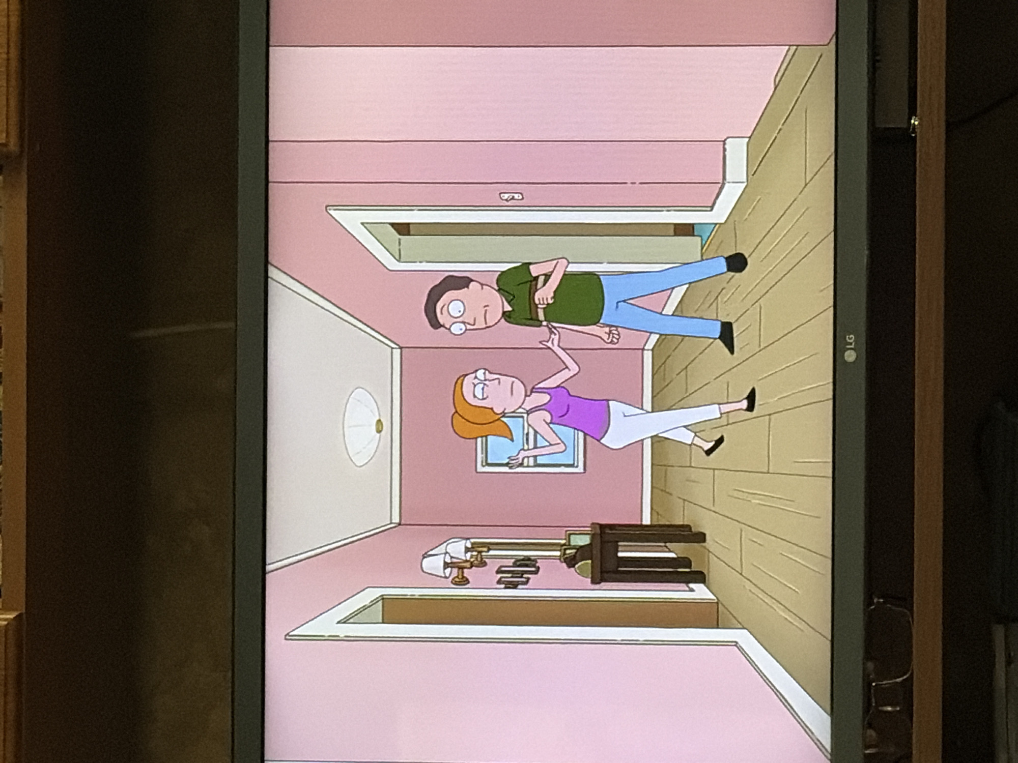 In the animated scene, a woman with orange hair in a purple tank top and white pants stands in a hallway with pink walls and a wooden floor. She is looking towards a man with brown hair wearing a green striped shirt and blue pants, who is standing near a doorway. The man is looking at the woman. A small table with a lamp and some objects is against the wall to the left. A window with blue panes is visible on the right wall. A circular light fixture is on the ceiling. The setting appears to be an interior of a residence, possibly a house.
