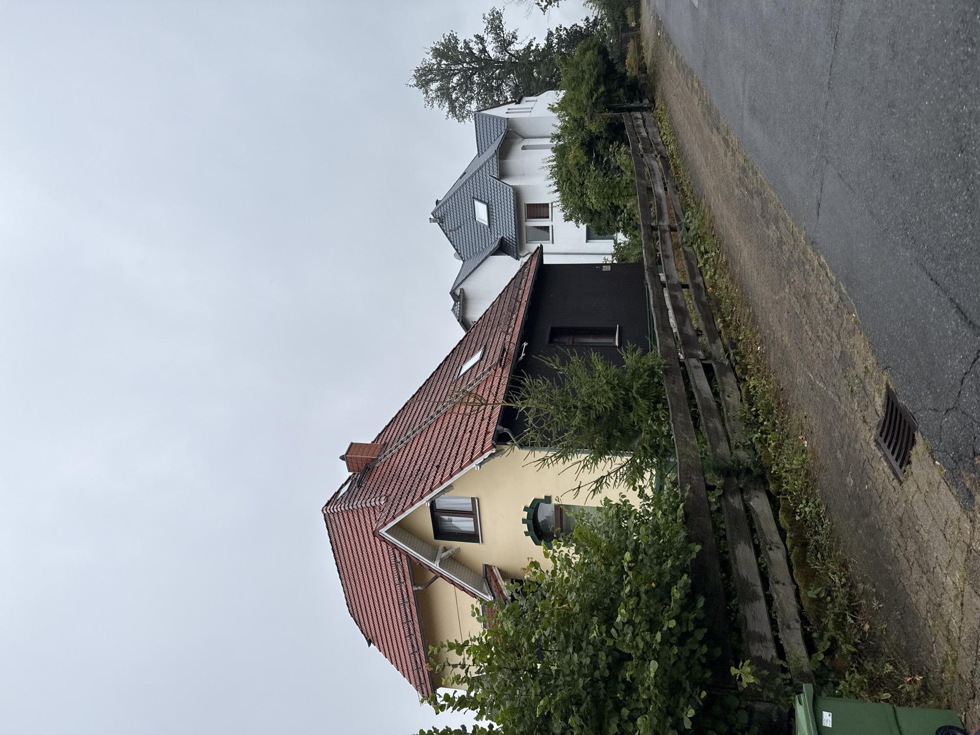 The image depicts a residential street in Bad Sachsa, Germany, under an overcast sky. A rustic wooden fence runs parallel to the street, separating a paved walkway from several houses. The first house visible is a two-story dwelling with a yellow facade and a red tile roof. Adjacent to it, a smaller, dark-colored structure is partially visible. Further down the street, a larger white house with a dark gray tile roof stands out. Lush green trees and bushes flank the houses and line the fence. A green trash bin is situated on the left side of the walkway, near a storm drain grate. The road surface is asphalt, showing some wear and tear. The scene is quiet and appears to be a typical residential area in a German town.