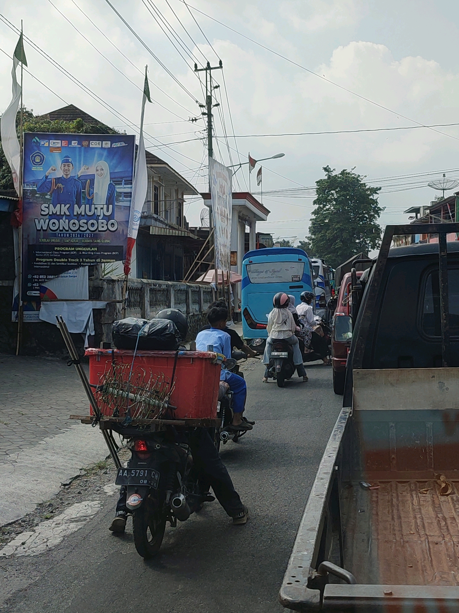 The image depicts a street scene in Kalikajar, Indonesia, with heavy traffic. In the foreground, a motorcycle is parked, carrying a large red container loaded with green, leafy plants. The motorcycle has a license plate that reads "AA 5791 CN". Behind this, several other vehicles are visible, including a blue bus with text on its rear, a red car, and a black pickup truck in the far right. People are seen riding motorcycles and walking on the side of the road. A large banner advertising "SMK MUTU WONOSOBO" is displayed on the left side of the street, featuring images of two young people in uniform. The environment is urban, with buildings lining the street and utility poles with numerous wires overhead. The sky is partly cloudy. The overall impression is one of a busy, active street in a town.