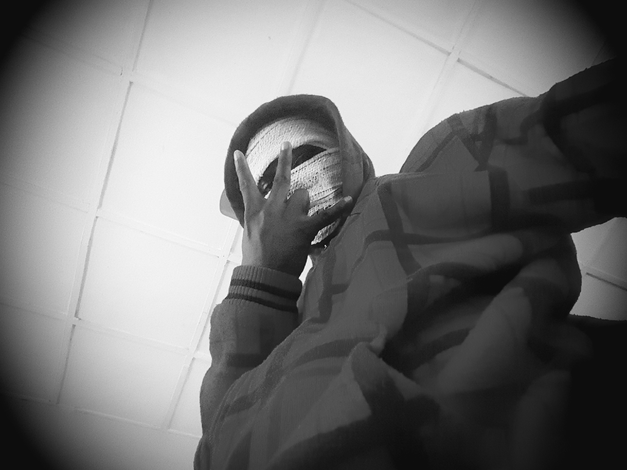 This black and white image, captured from a low angle, depicts an individual from the chest upwards. The person's head is covered by a hooded garment, under which a light-colored, textured head covering is visible. The face is not discernible. The individual's right hand is raised, forming a "V" gesture with the index and middle fingers extended, while the other fingers are curled inward. The sleeve of the garment shows three horizontal stripes near the wrist. The main body of the clothing features a subtle, darker patterned design. The background consists of a brightly lit, light-colored ceiling structured with a grid pattern of lines, indicative of ceiling panels or tiles. A strong vignette effect darkens the periphery of the image.