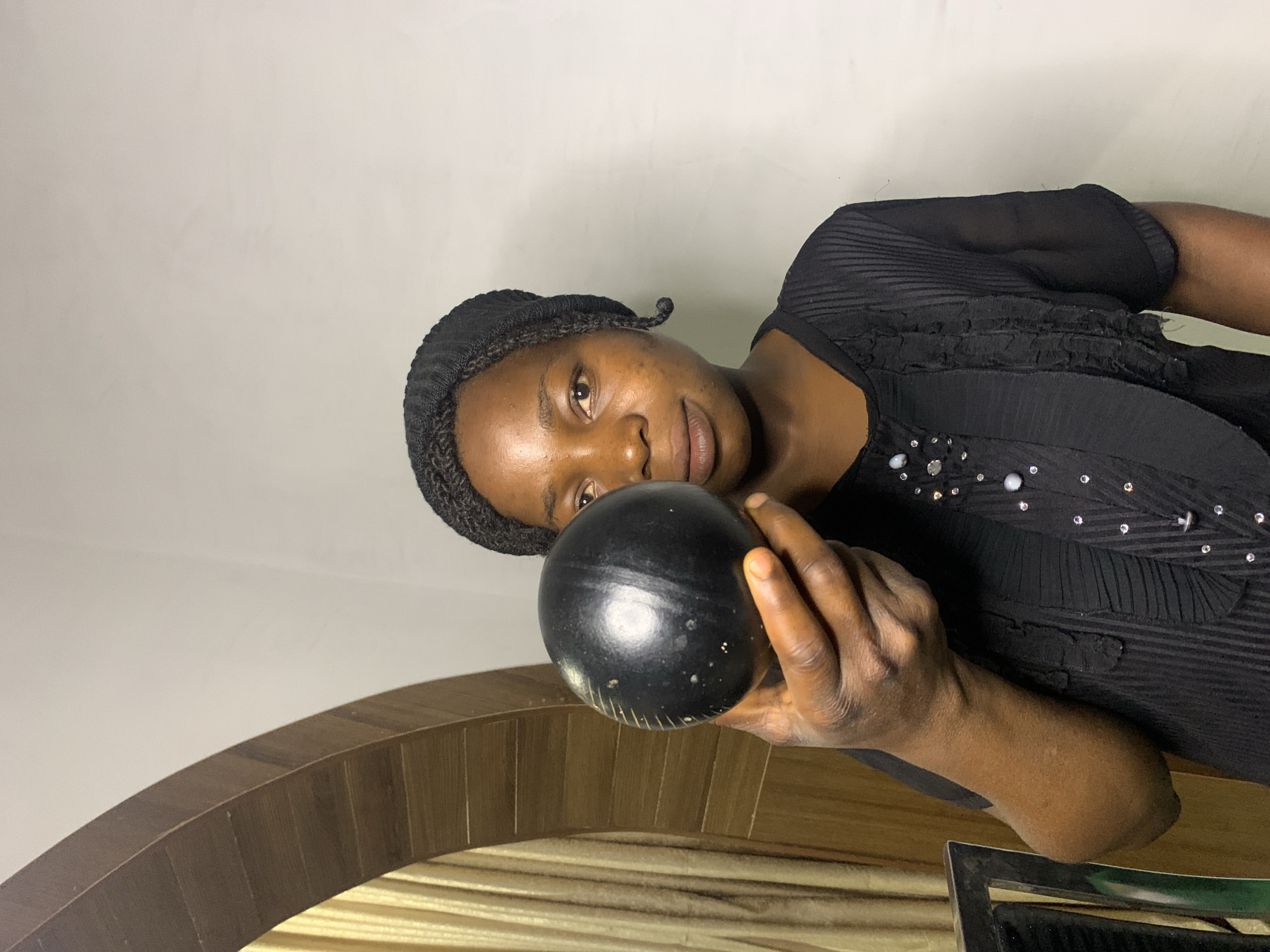 A woman is holding a black, spherical object in front of her face. She is wearing a black head covering and a black, ruffled shirt. She is standing in what appears to be a room with a light-colored wall and a wooden archway in the background. The scene is indoors, likely in a residential setting in Jos, Nigeria.