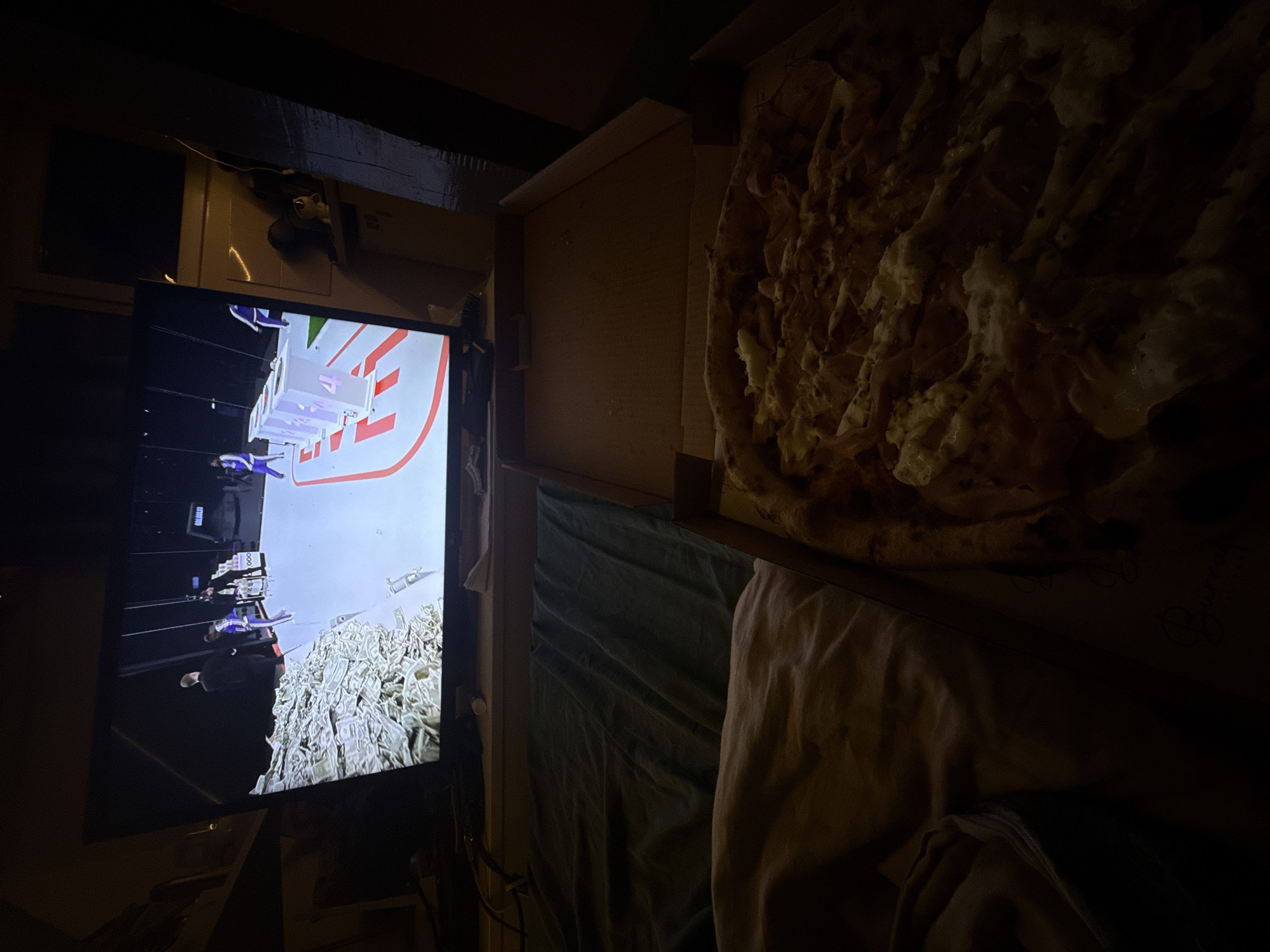 A dimly lit interior shows a television screen on the left displaying a scene from what appears to be a heist movie or video game involving money. To the right of the screen is an open pizza box resting on bedding. The pizza is topped with cheese and what appears to be dough. The image suggests a casual setting, possibly a bedroom or living room, where someone is watching TV and eating pizza. There is no information provided in the image regarding the location being in Sofia, Bulgaria.