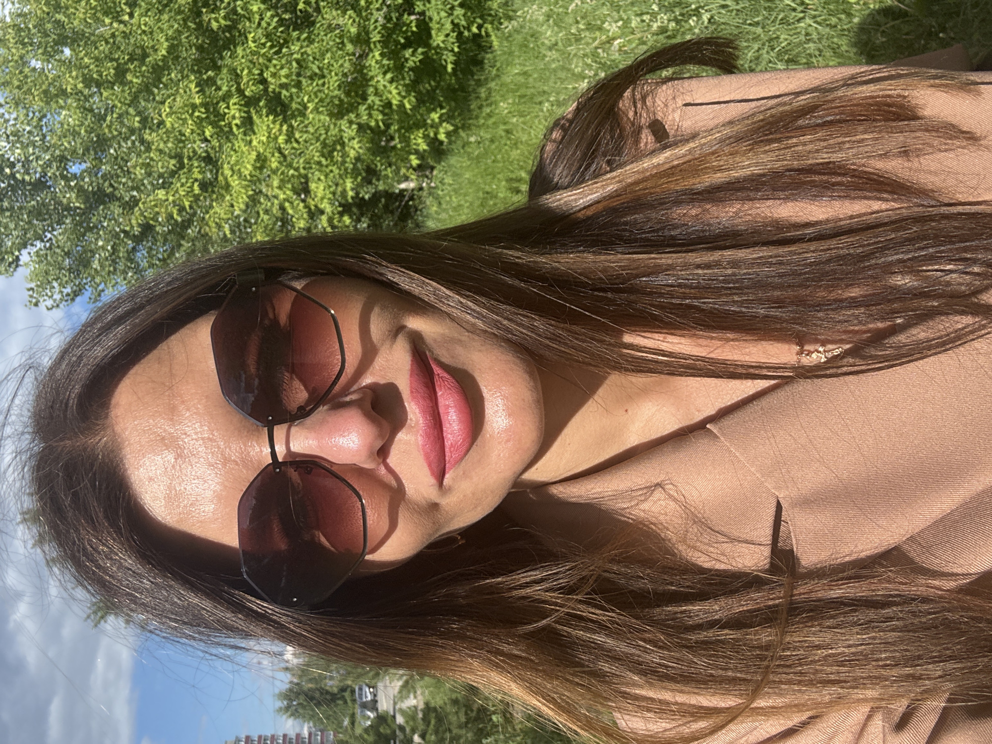 A medium shot shows a woman with long, brown hair and pink lipstick, wearing sunglasses and a tan blazer, outdoors. The woman is looking at the camera. Behind her, a green tree and grass are visible, as well as a blue sky with clouds. In the distance, there are buildings and cars, suggesting an urban environment. The lighting indicates it is daytime and sunny. The location is identified as Chaikovsky, Russia.
